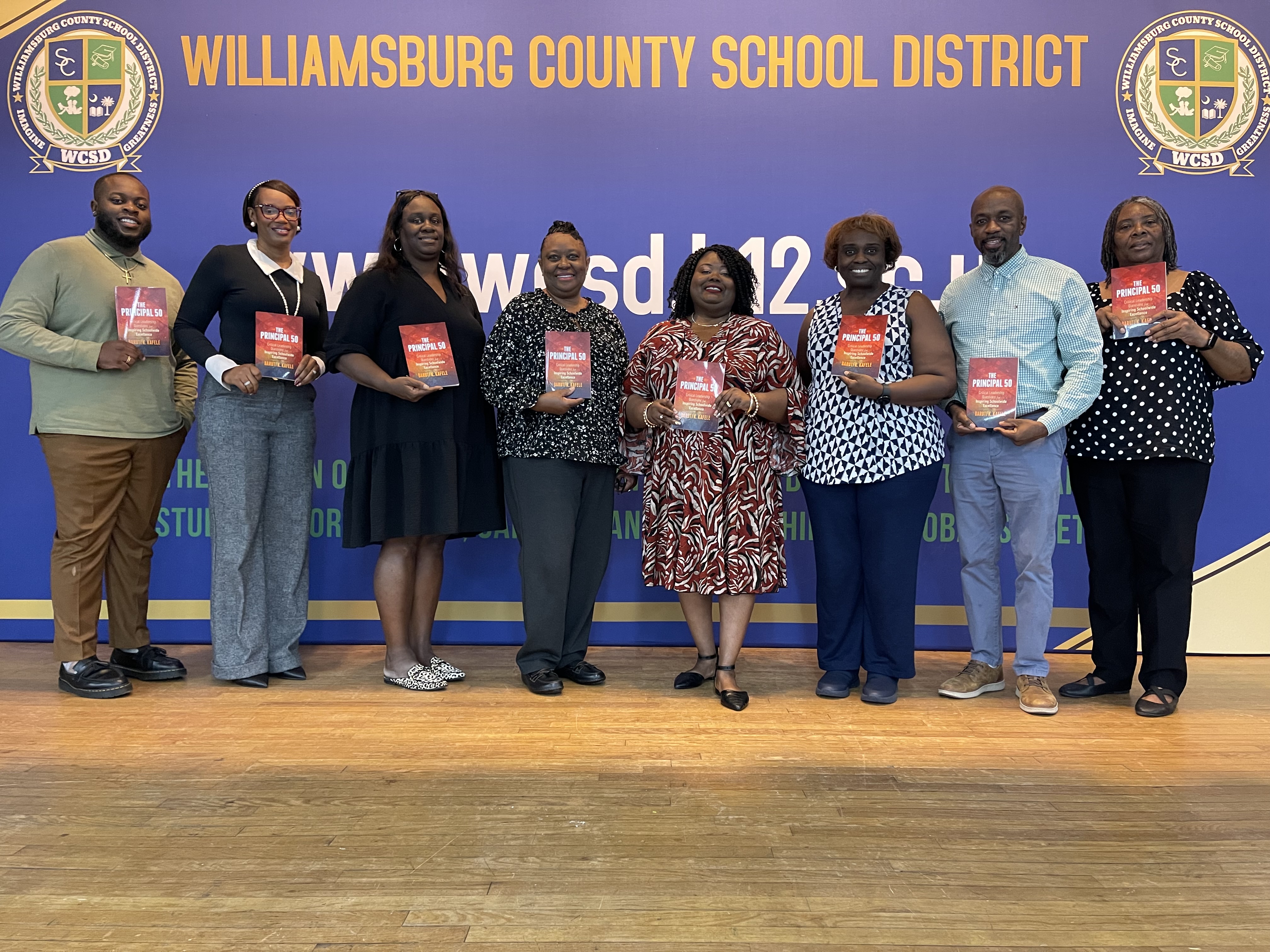 Eight Williamsburg County School District staff members stand together in front of a blue and gold district backdrop, smiling and holding copies of *The Principal 50*. The Williamsburg County School District name and WCSD logos are prominently displayed behind the group.