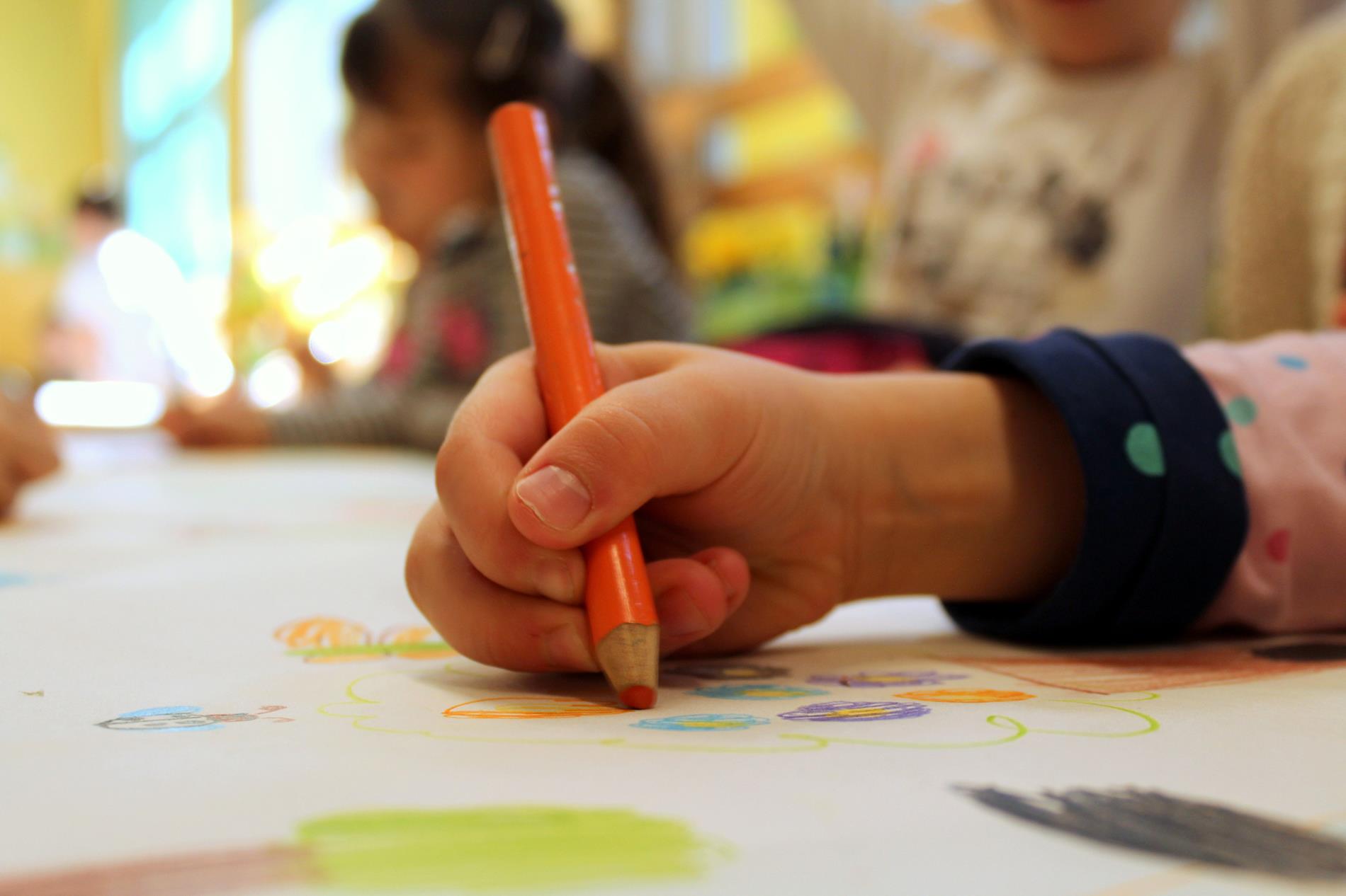 A small child's hand holds and colors with an orange colored pencil