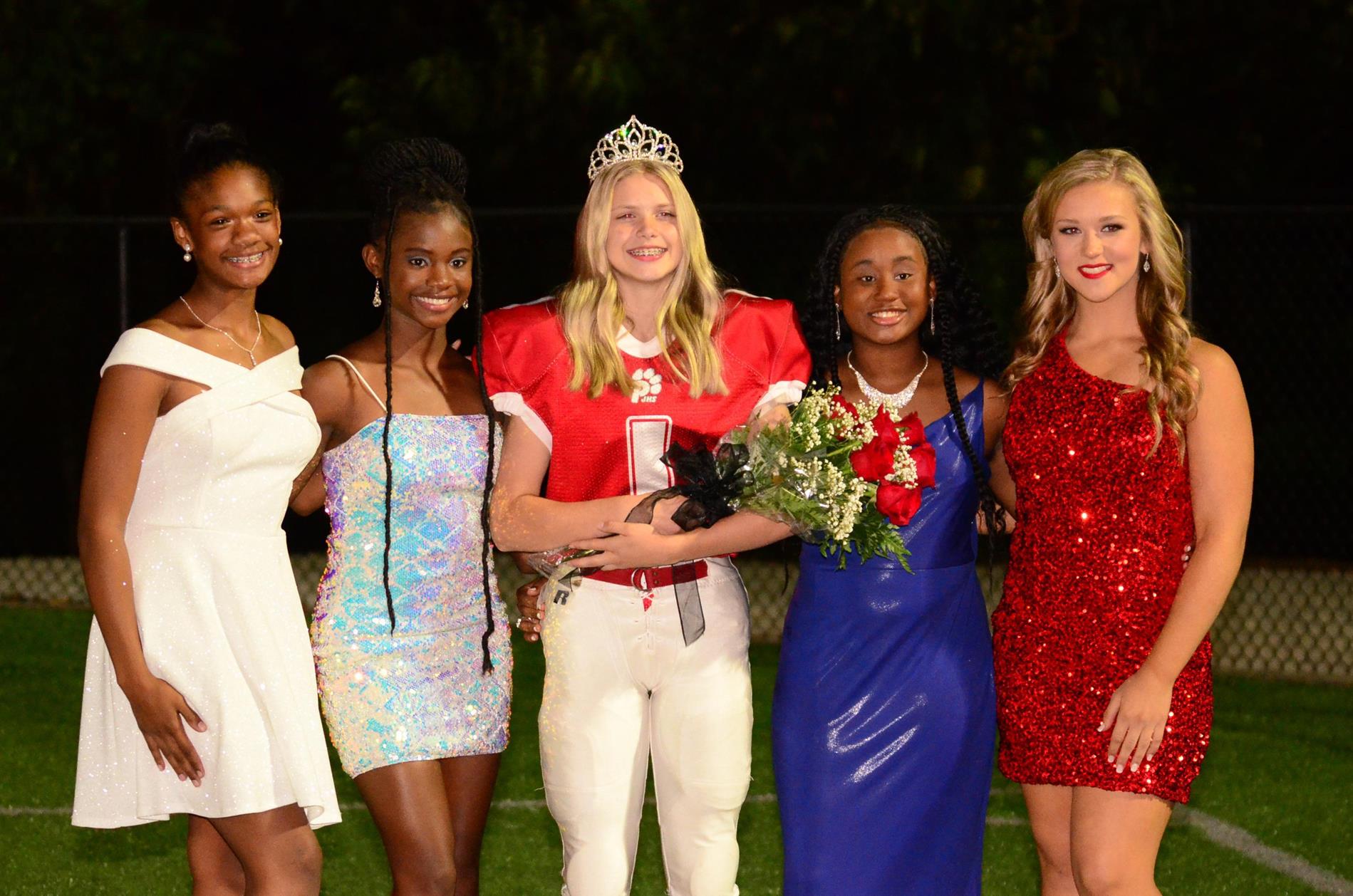 Homecoming queen, Ann Hartley, pictured with court. Girls are wearing formals with the exception of Ann who is wearing her football uniform and crown while holding flowers