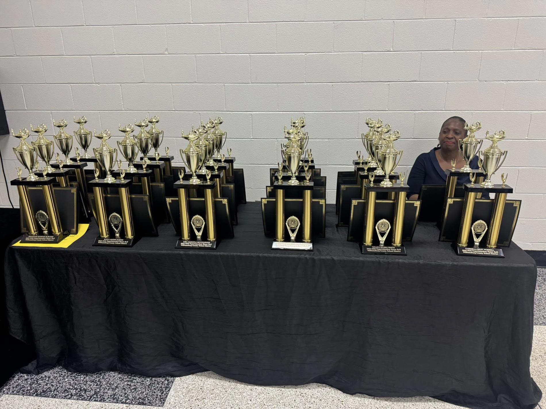 table with trophies on it. lady is seated behind the table 