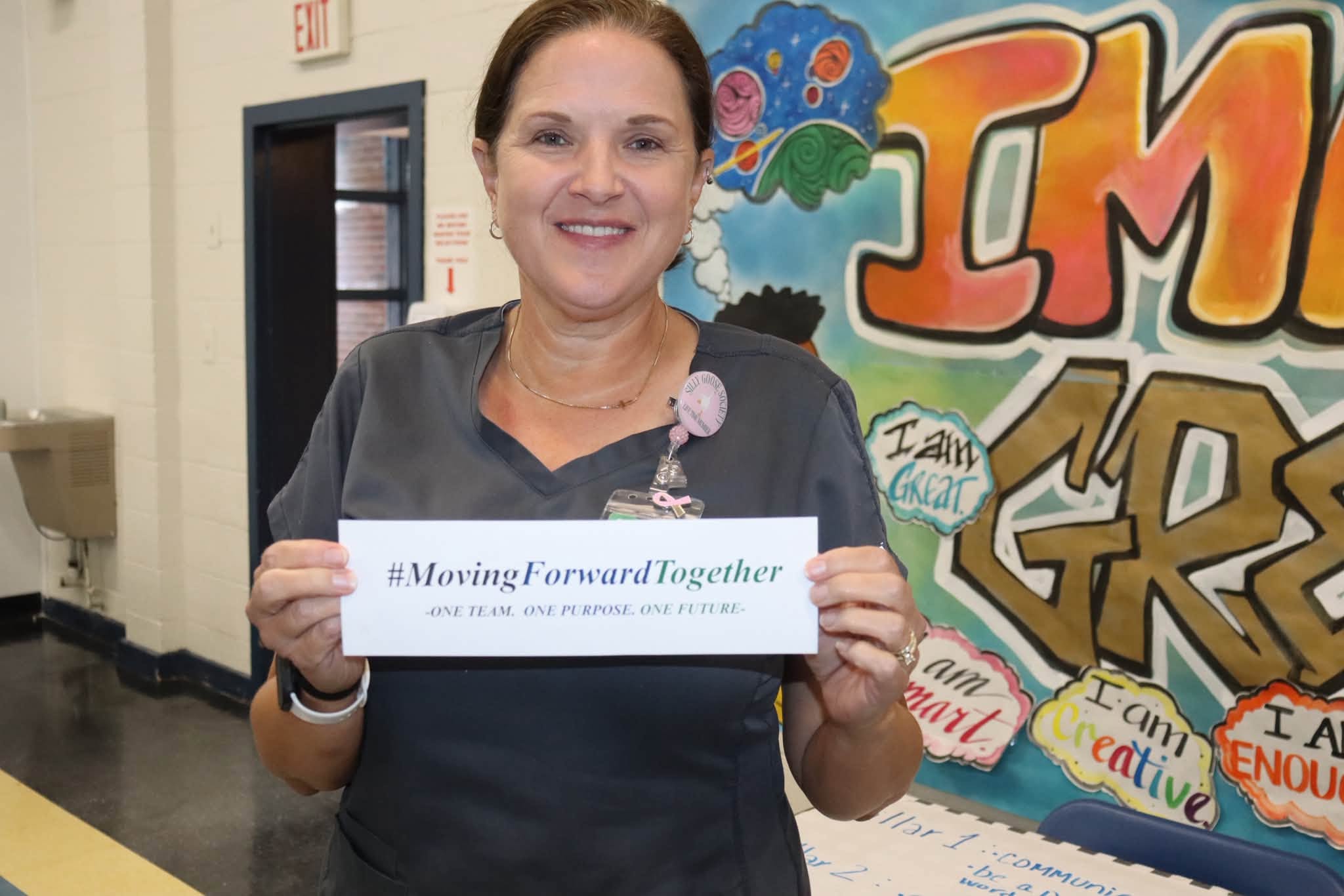 A smiling Williamsburg County School District staff member wearing dark gray scrubs holds a card reading “#MovingForwardTogether – One Team. One Purpose. One Future.” She stands in front of a colorful educational mural during a Summer Leadership event.