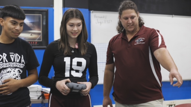 A male teacher stands with one male and one female student giving instruction 