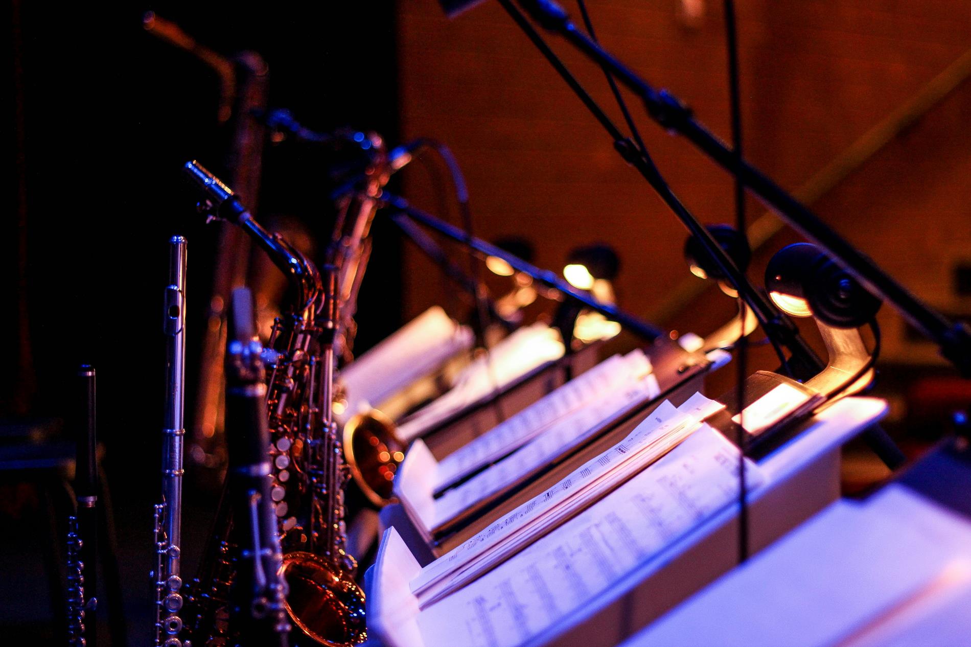 A group of music stands with sheet music and flutes, saxphones, and other instruments sit in low light
