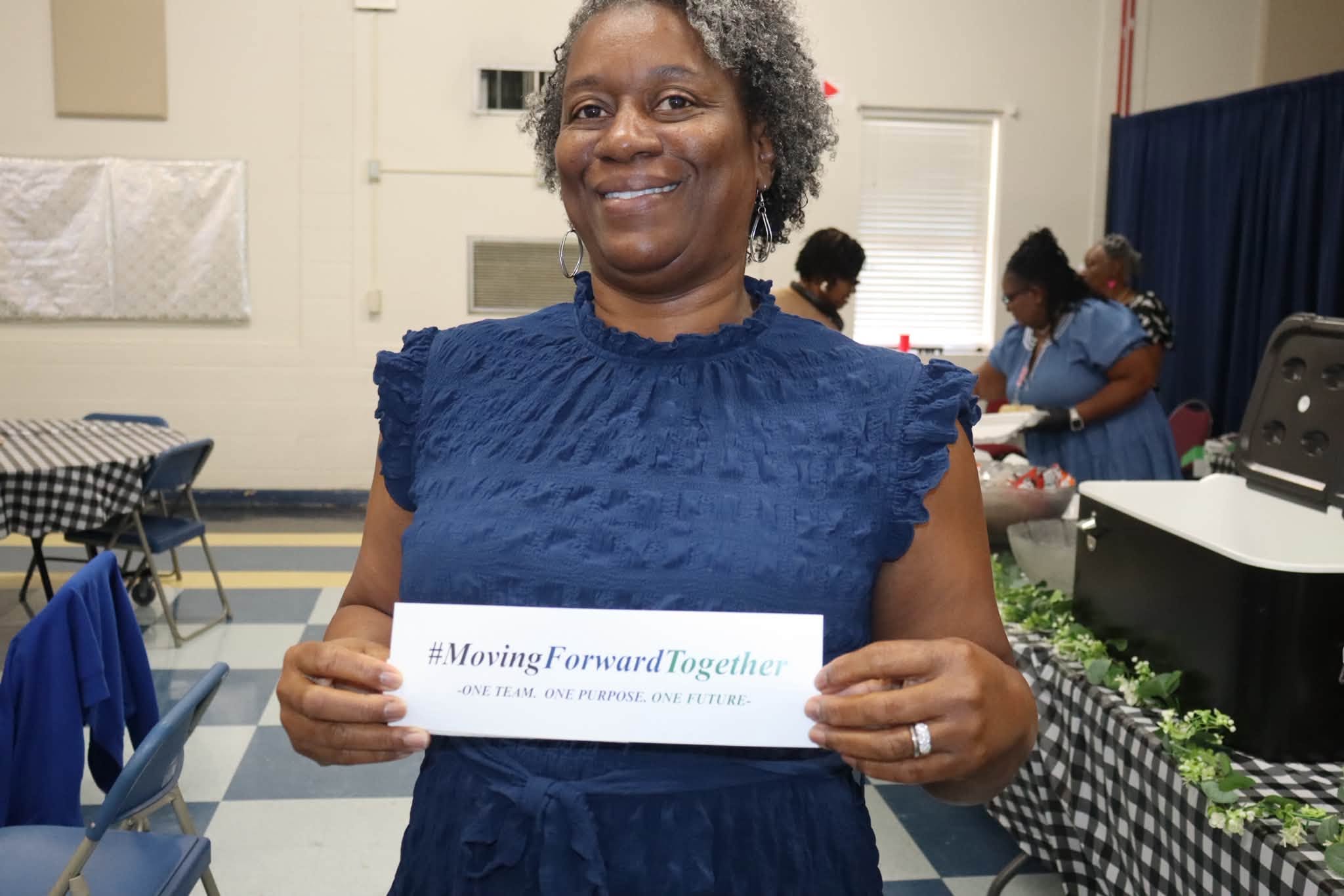 A smiling Williamsburg County School District staff member wearing a navy blue dress holds a card reading “#MovingForwardTogether – One Team. One Purpose. One Future.” She stands in the event space during a Summer Leadership session, with tables and other attendees visible in the background.