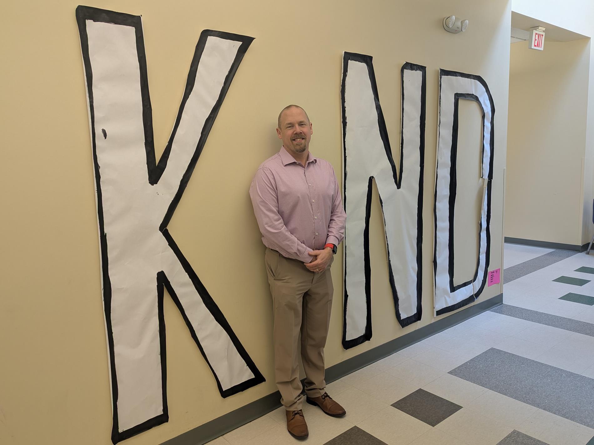 Picture of Executive Director wearing a pink shirt. The Director stands agains a the wall, forming the "i" in a large sign reading "KIND".