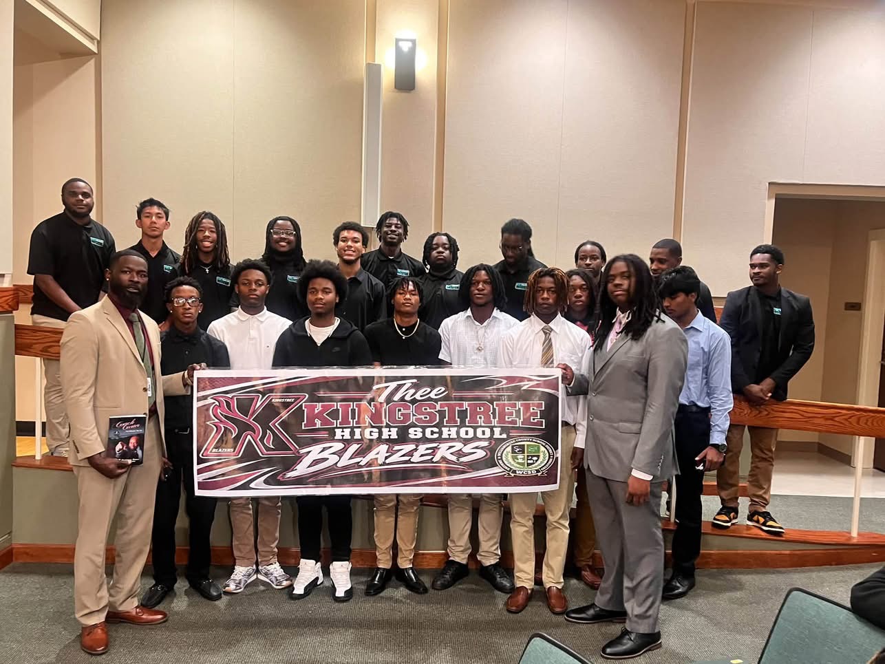 A group of students and adults pose indoors while holding a “Thee Kingstree High School Blazers” banner.