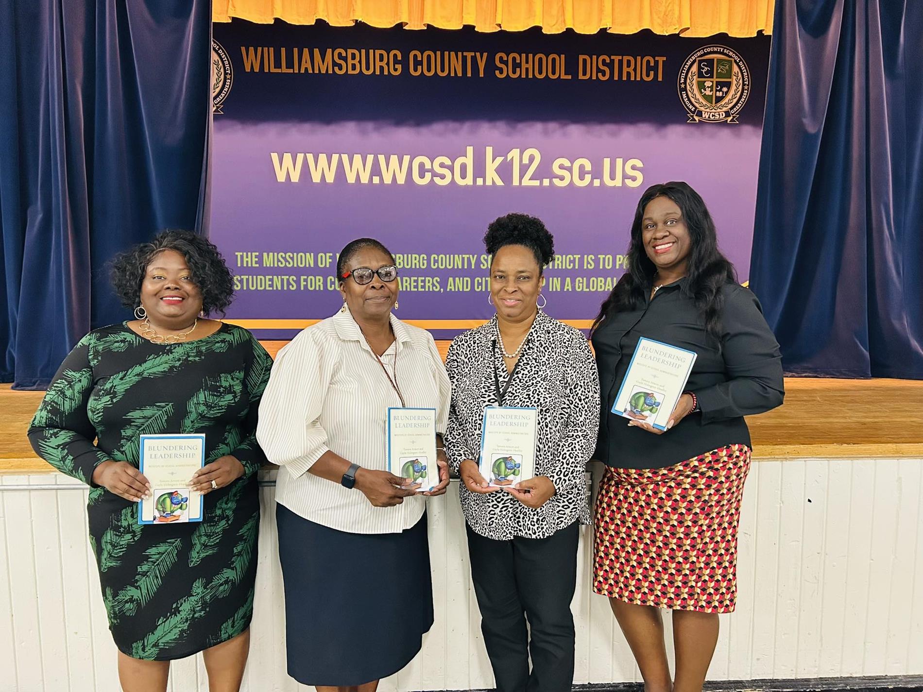A group of nine adults stand in a row holding matching books in front of a stage backdrop that reads "Williamsburg County School District" and "www.wcsd.k12.sc.us