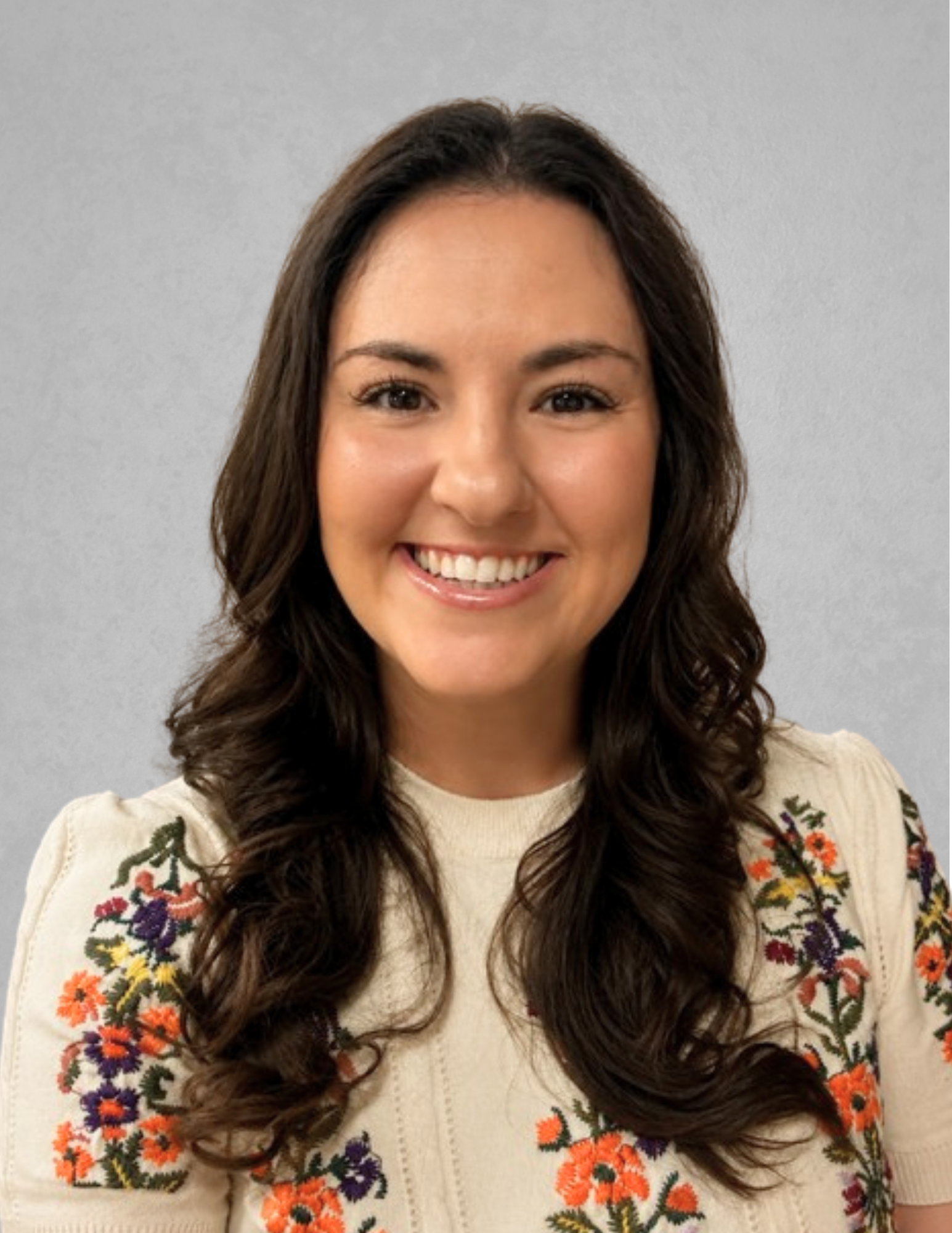 Professional headshot of McKenzie Bienvenu smiling and facing the camera. She is wearing a cream-colored blouse with colorful floral embroidery on the sleeves and shoulders. Bienvenu has long dark wavy hair and is posed against a light gray background.