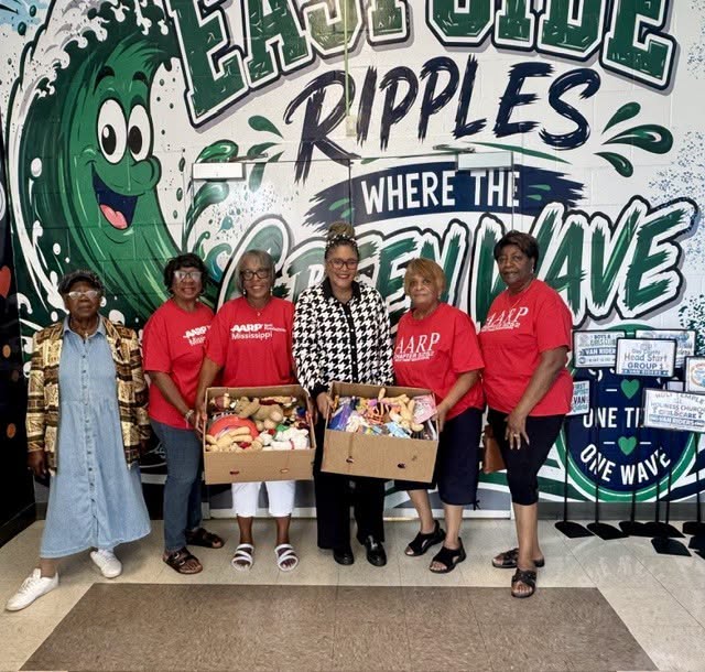 AARP representatives and an East Side Elementary staff member pose with boxes of donated items for the school’s PBIS store in front of the East Side Ripples mural.