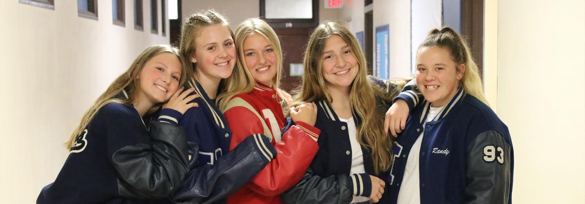 5 teenage girls posing in a hallway wearing letterman jackets.