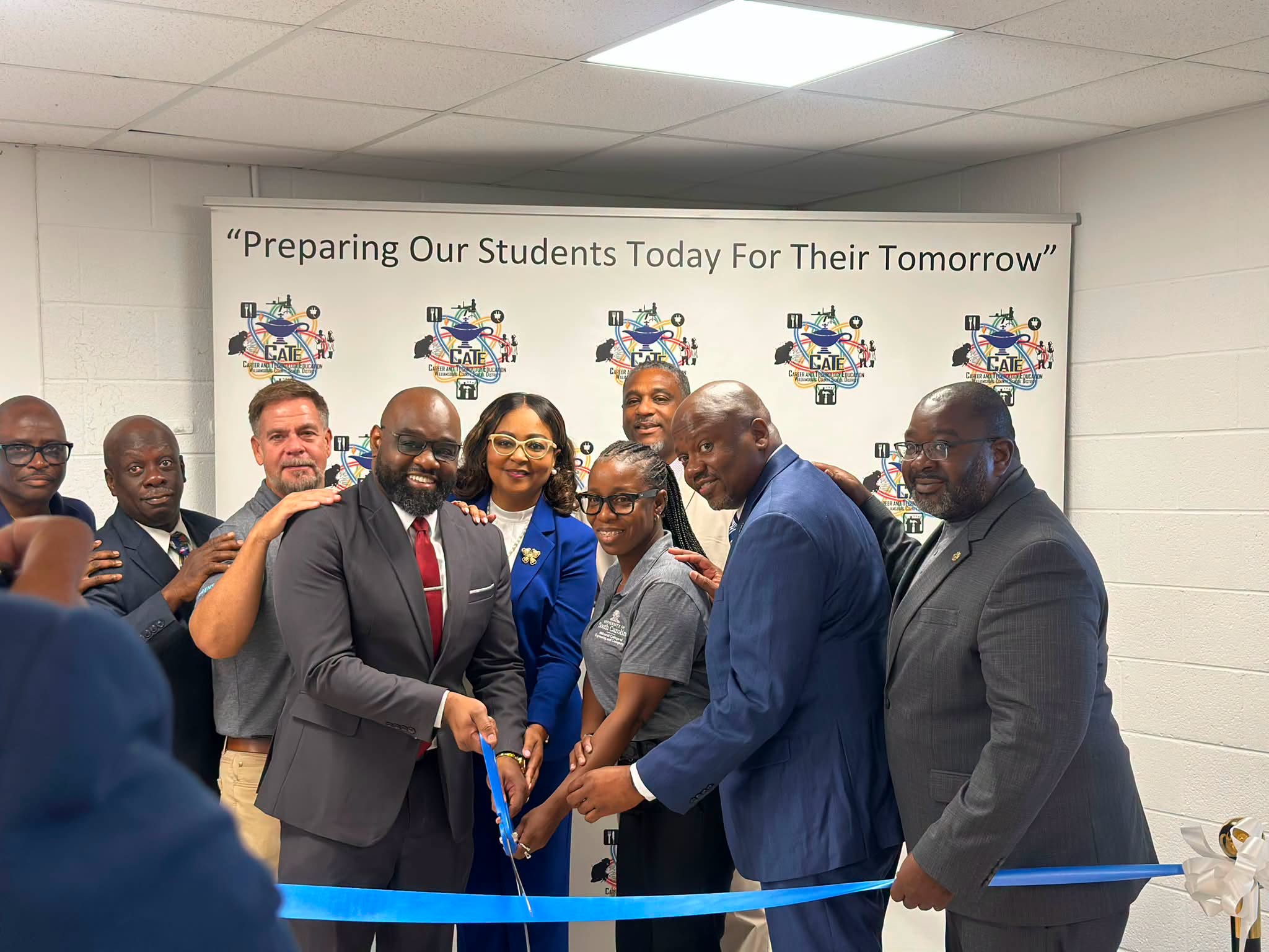 A group of school and community leaders gathers for a ceremonial ribbon cutting, smiling and posing behind a bright blue ribbon. Several participants hold scissors at the center. Behind them is a Career and Technology Education (CATE) backdrop featuring the message, “Preparing Our Students Today For Their Tomorrow.”