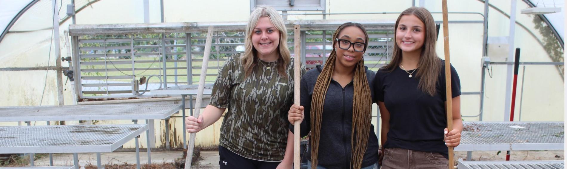 Three girls holding shovels inside the school greenhouse
