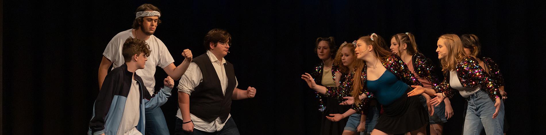 Two groups of students face each other on stage in a theater production. They appear to be dancing.