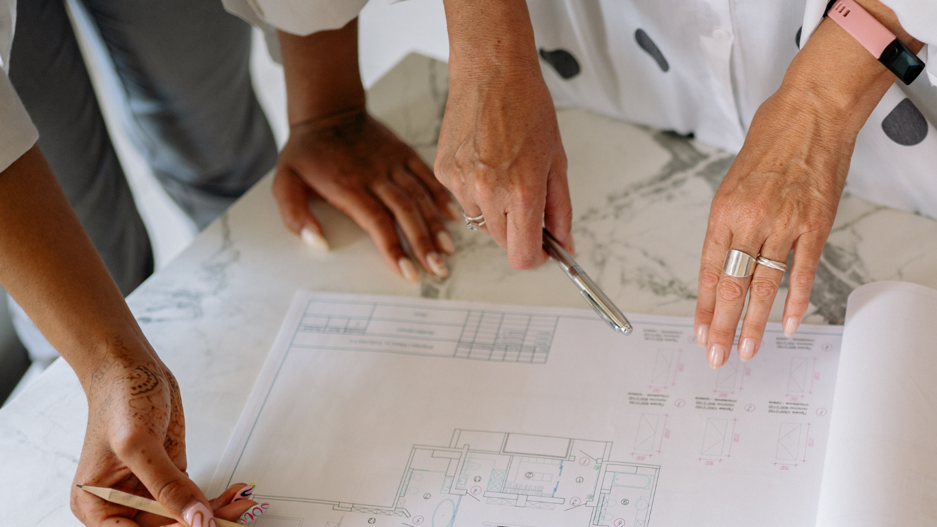 Two women's hands holding pens gesture over a blueprint of designs on a marble countertop