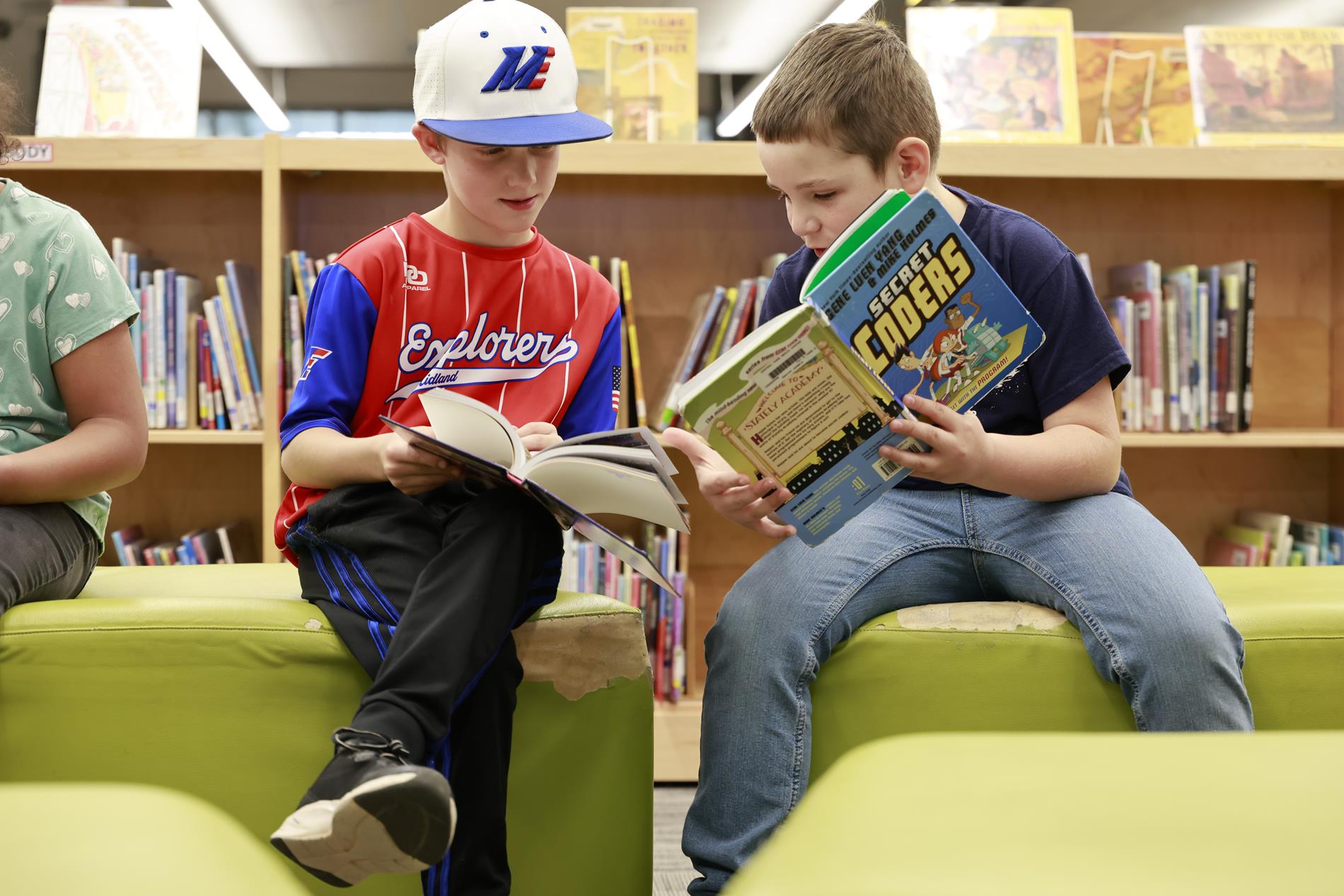 Two elementary aged boys sit in the Central Park library, each sharing a book they are reading with one another