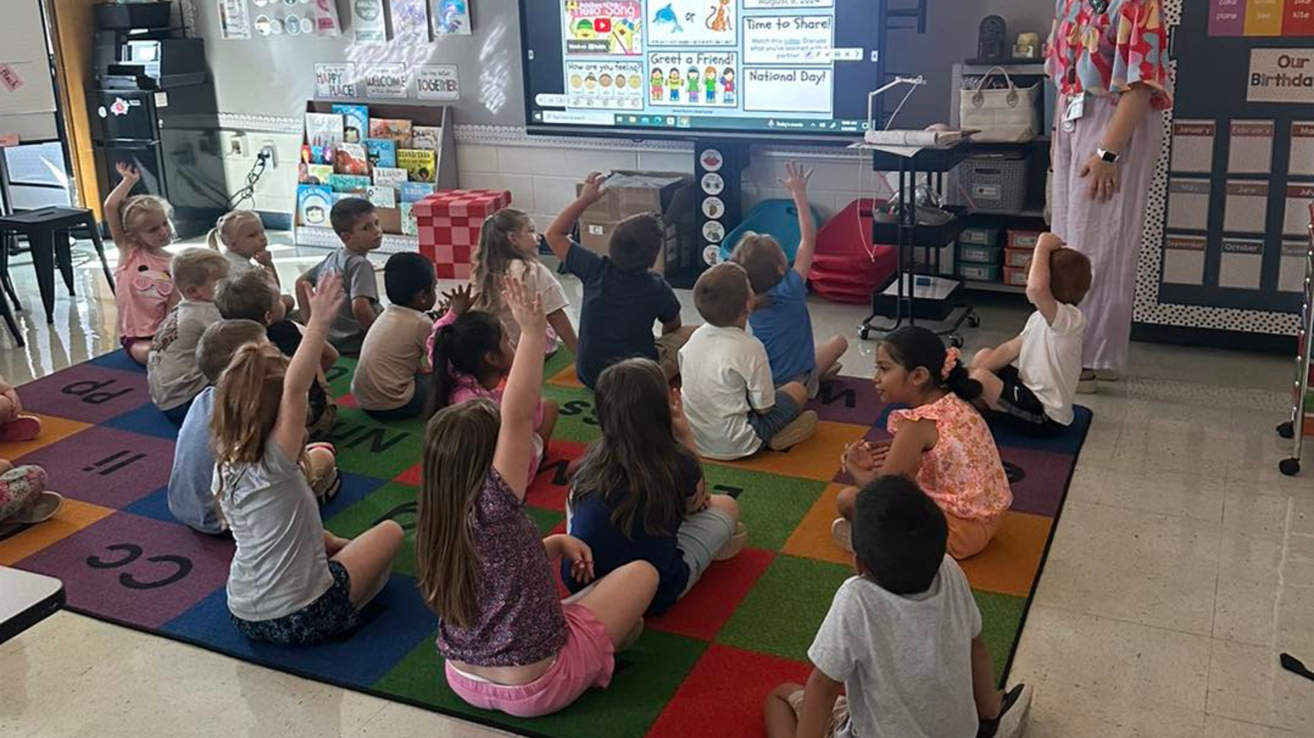 Students are all sitting on the rug and looking towards their teacher. Some are raising their hand to answer a question.