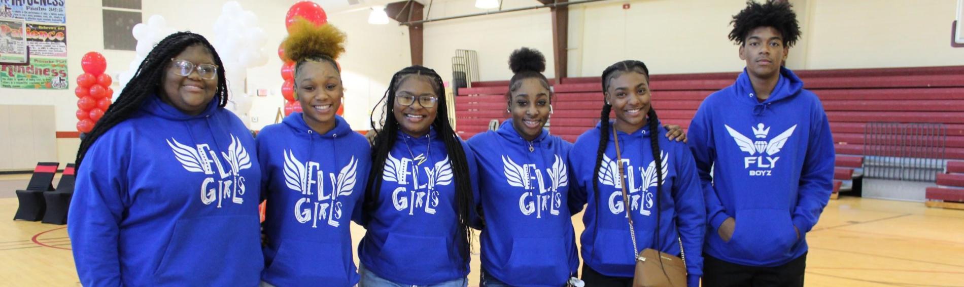 Five girls and a boy pose in the GWCFC gym wearing matching blue hoodies that say FLY girls and FLY boy 
