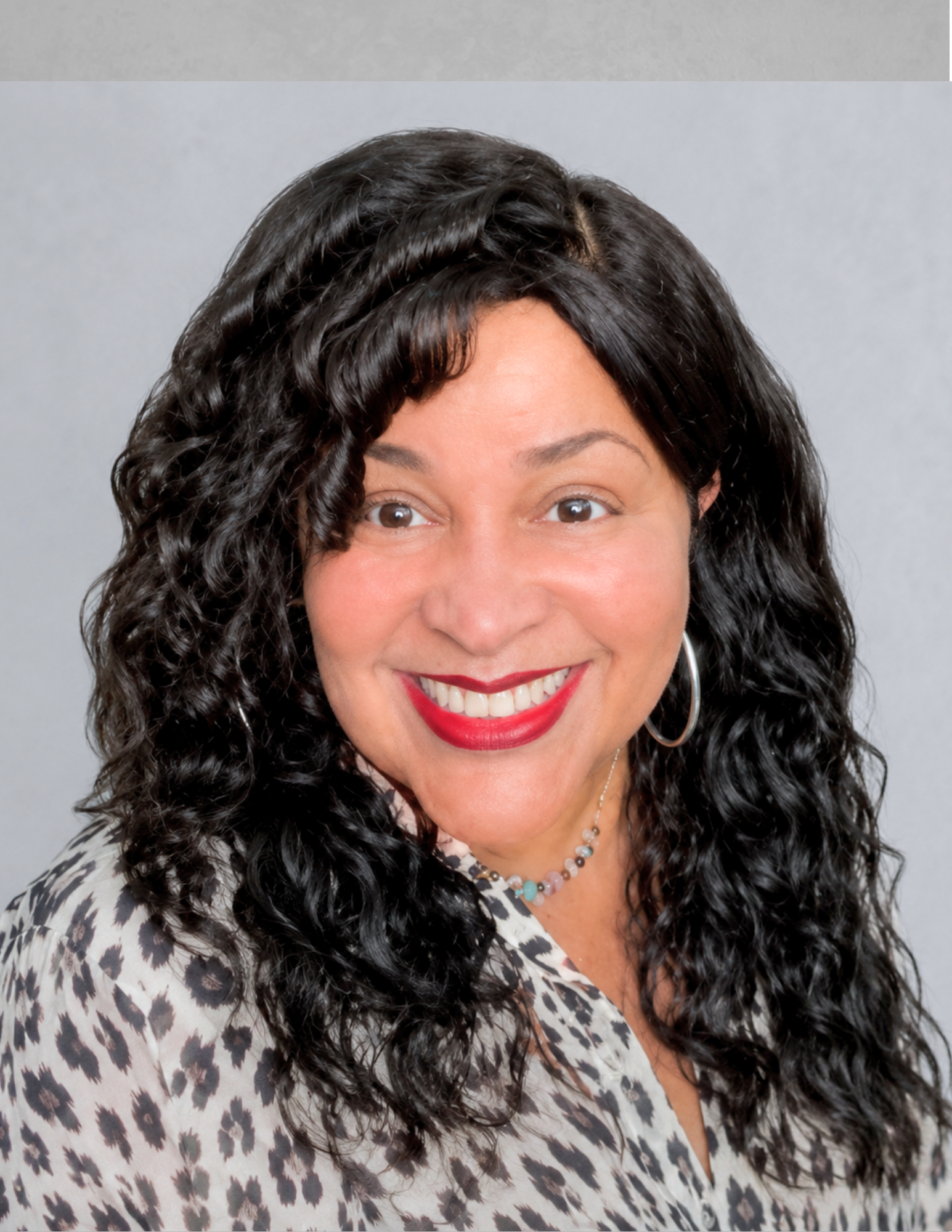 Professional headshot of Valerie Bandy smiling and facing the camera. She has long dark curly hair, red lipstick, and is wearing silver hoop earrings, a beaded necklace, and a patterned blouse. The portrait is set against a light gray studio-style background.