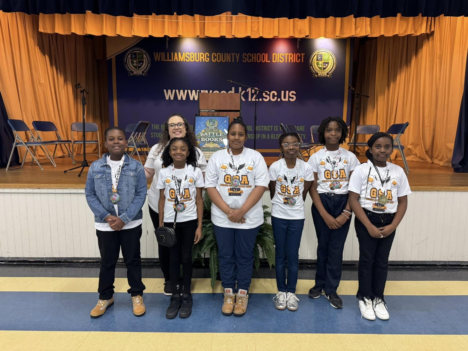 students and one adult wearing graphic tshirts in front of a blue and orange curtained backdrop with a banner that reads "WILLIAMSBURG COUNTY SCHOOL DISTRICT" and the district's website, "www.wcsd.k12.sc.us." 