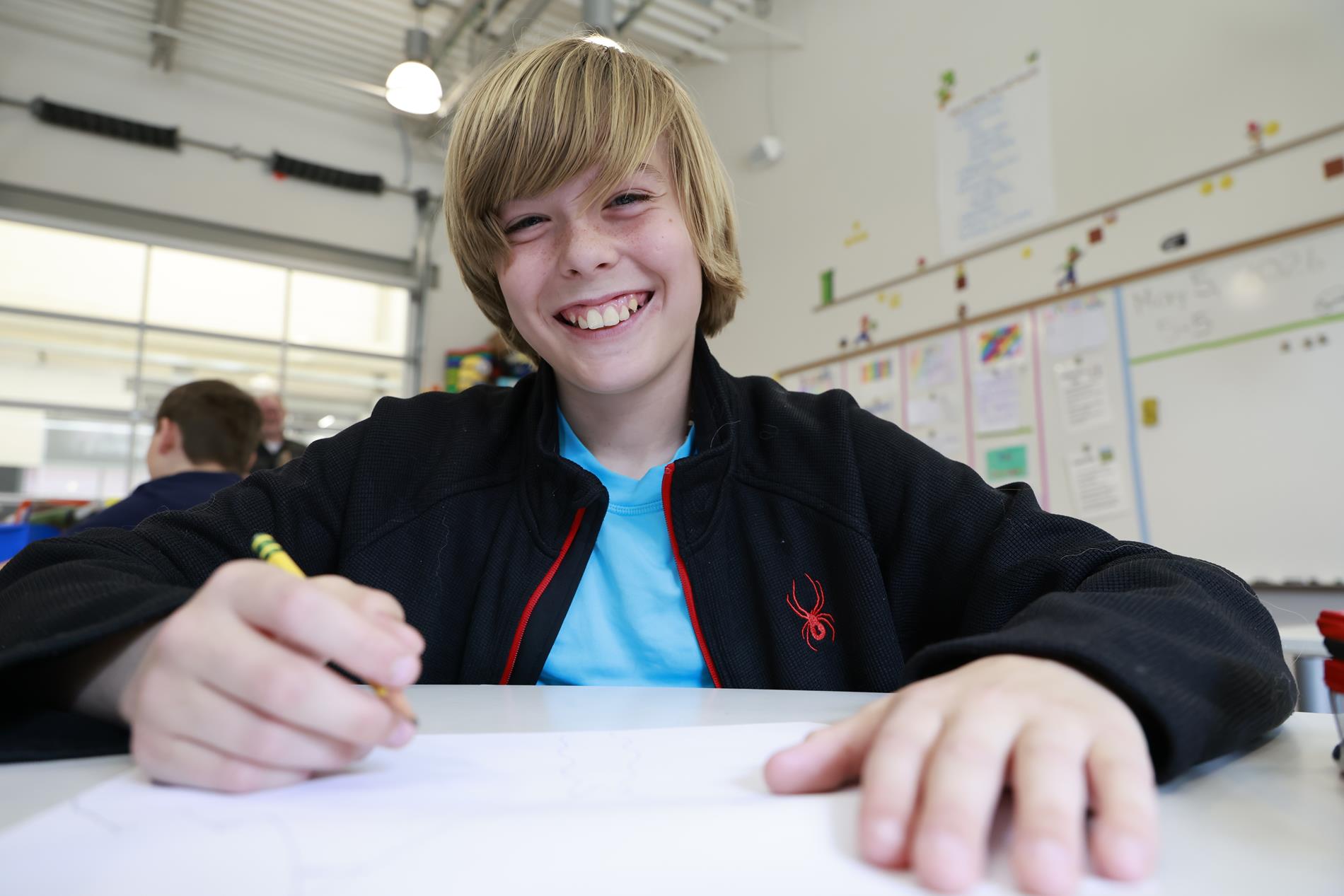 A young boy in 4th grade sits at his desk holding a penci, smiling at the camera