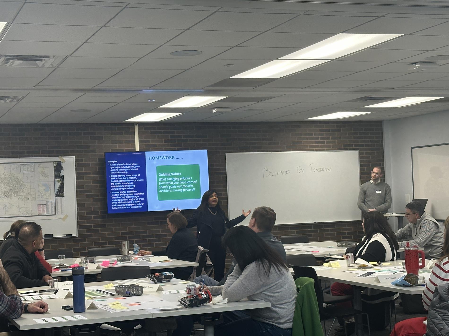 Steering Team members sit amongst tables in a conference room listening to a presentation