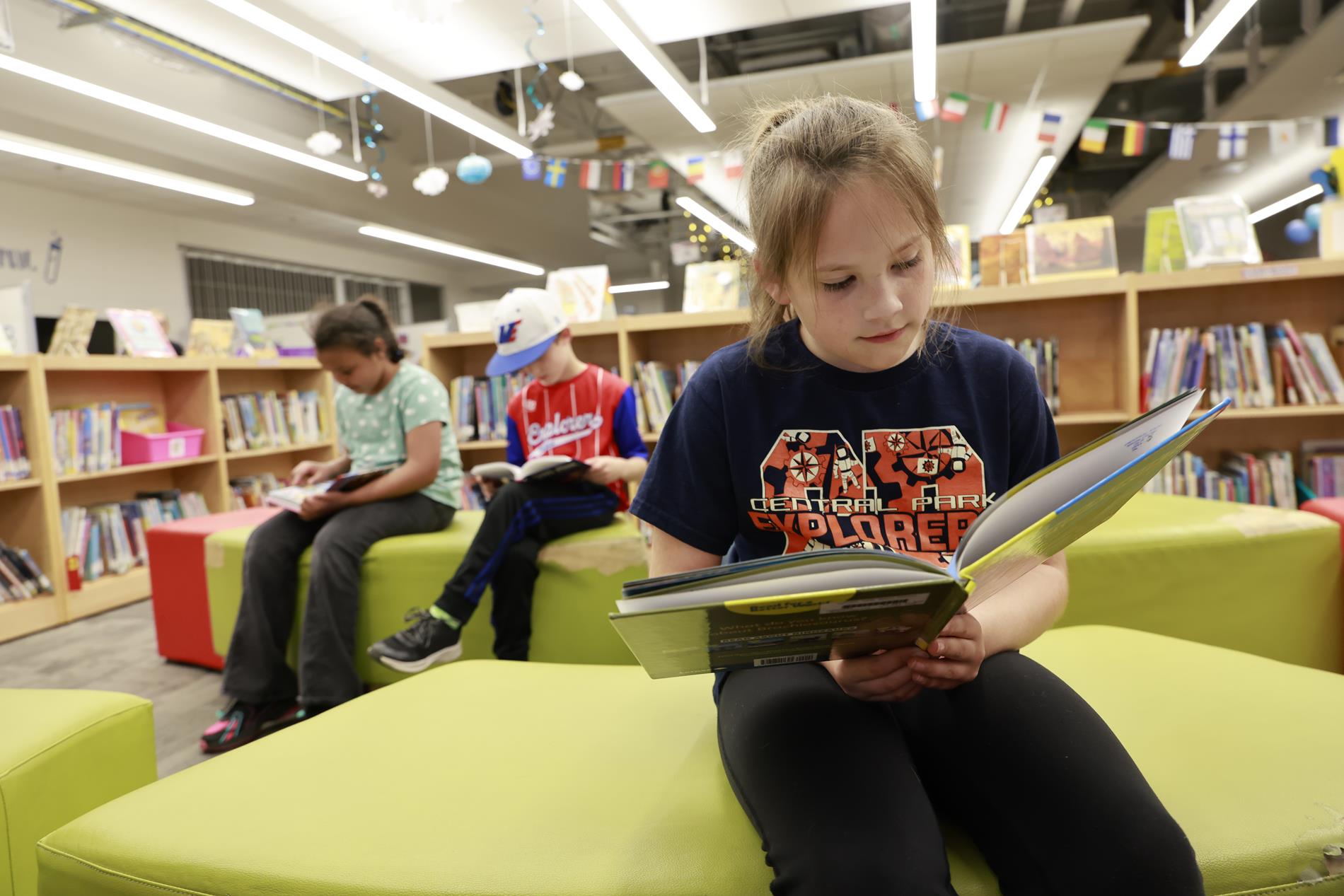 A third grade girl sits on a brightly colored sofa in the media center while reading a book with two other kids in tha background also reading