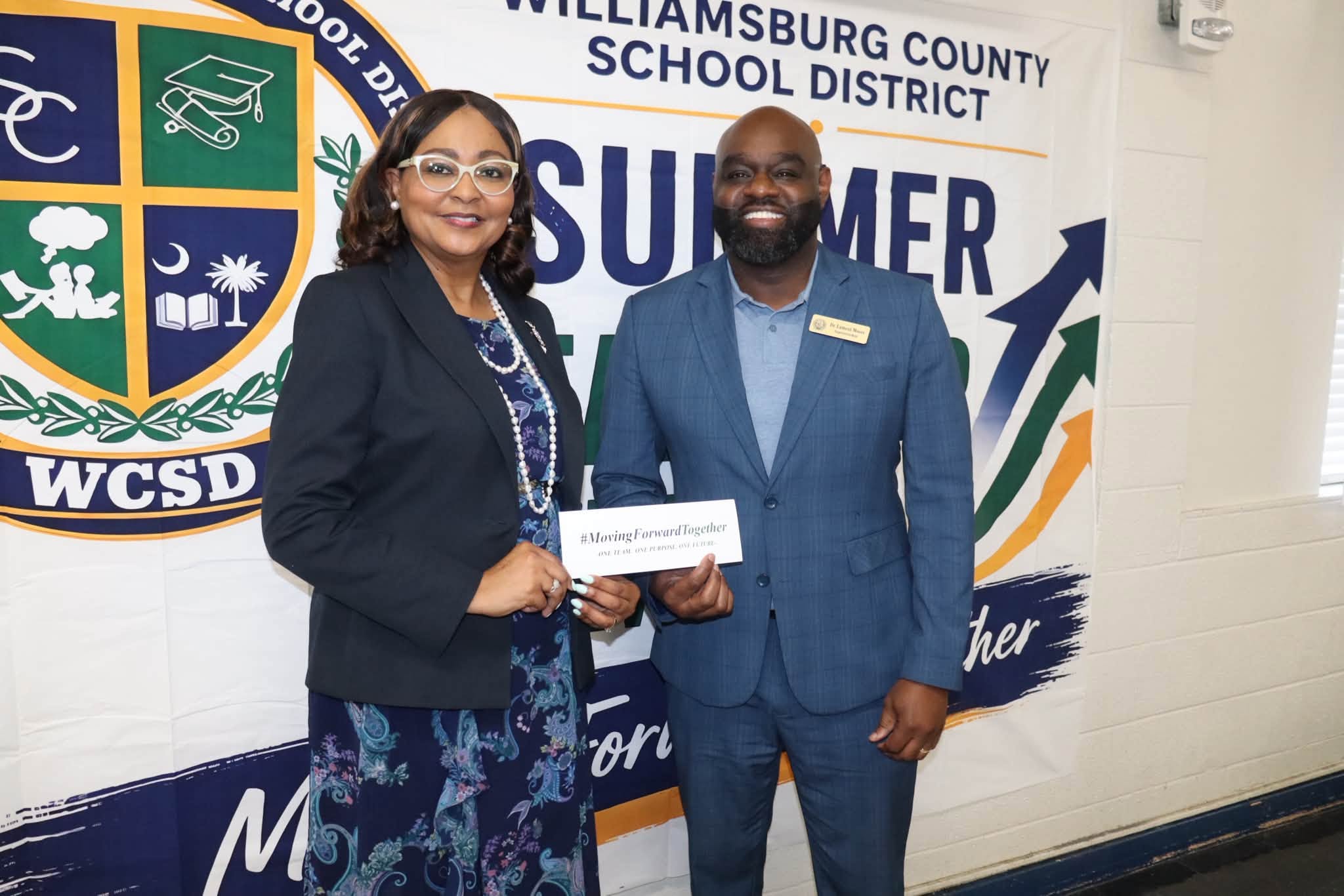  Two professionally dressed Williamsburg County School District staff members stand smiling in front of a large WCSD Summer Leadership backdrop. They are holding a card together that reads “#MovingForwardTogether,” with the district logo and “Summer Leadership” displayed prominently behind them.