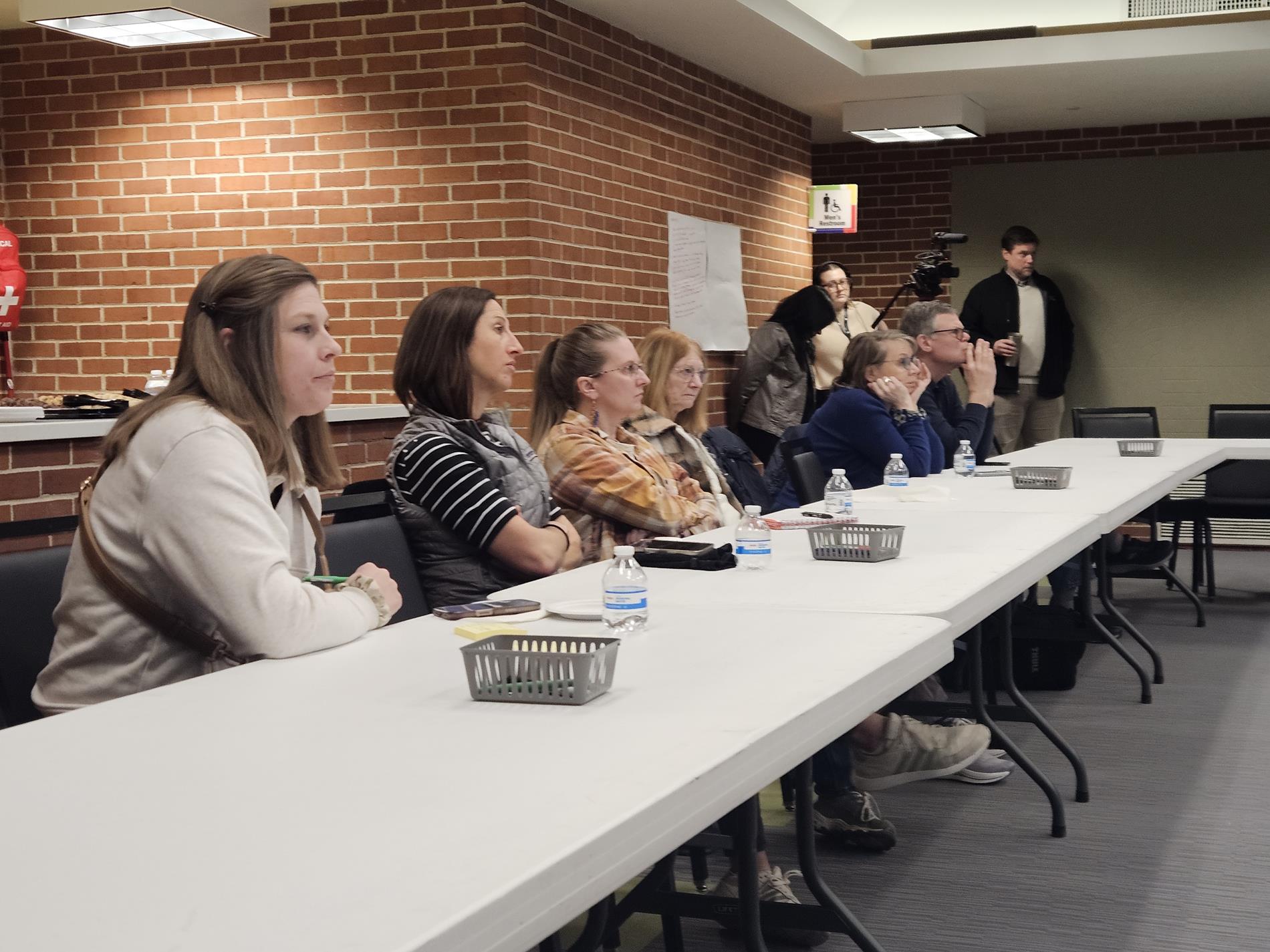 A group of adult attendees sit at tables in the Community Room listening to the presentation