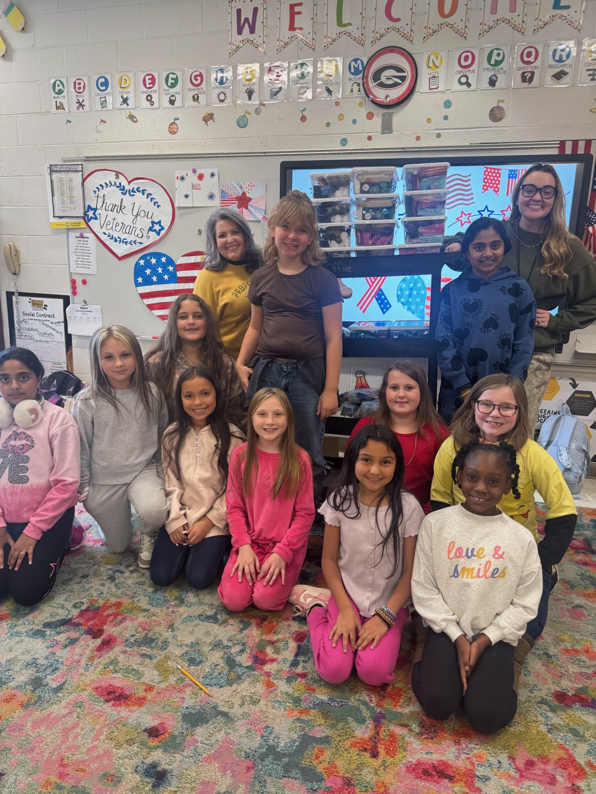 A group of smiling elementary students and two adults pose together in a colorful classroom. Patriotic student artwork and a “Thank You Veterans” display decorate the area behind the group.
