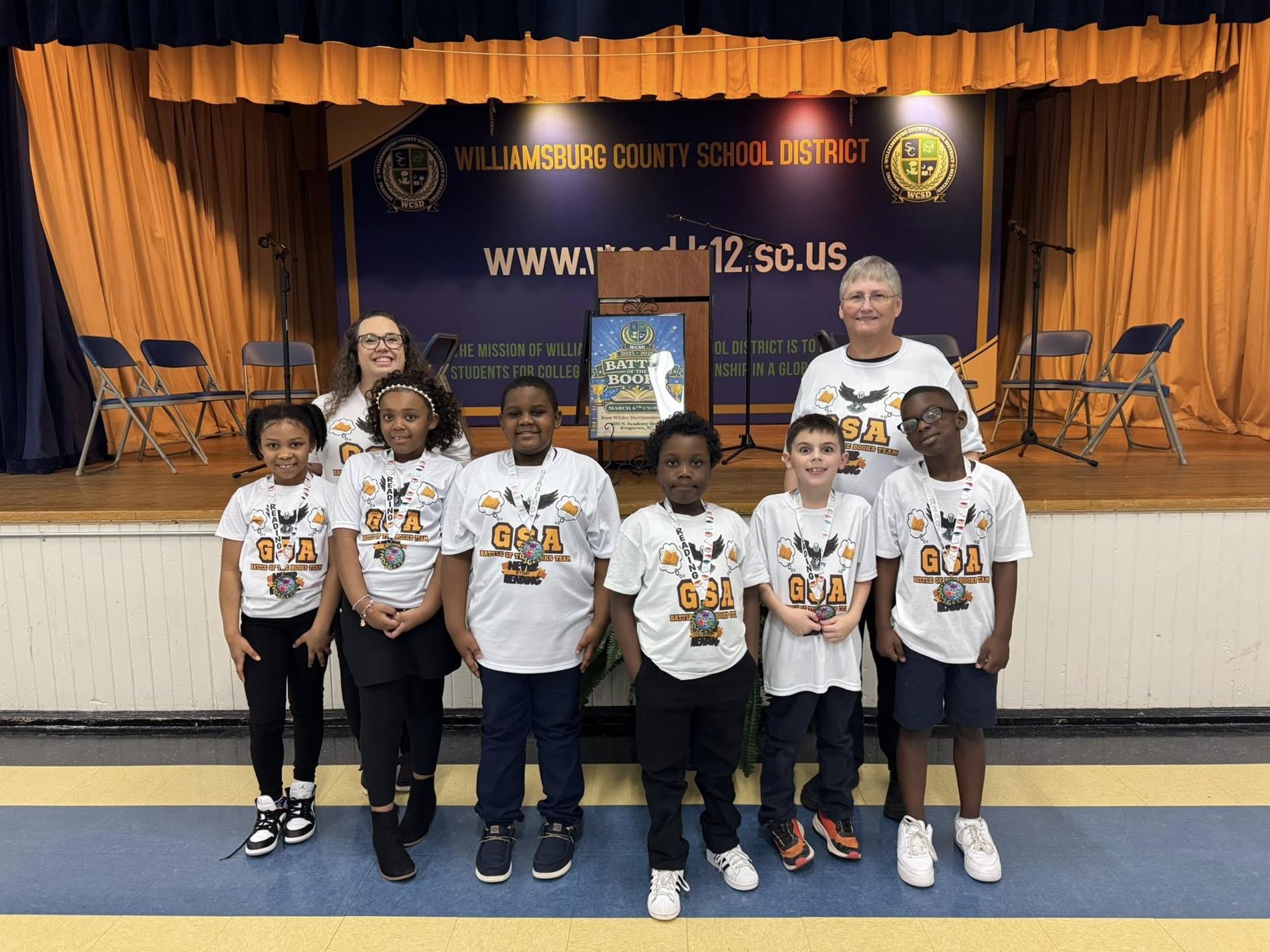 two female adults and small children in front of a blue and orange curtained backdrop with a banner that reads "WILLIAMSBURG COUNTY SCHOOL DISTRICT" and the district's website, "www.wcsd.k12.sc.us." 