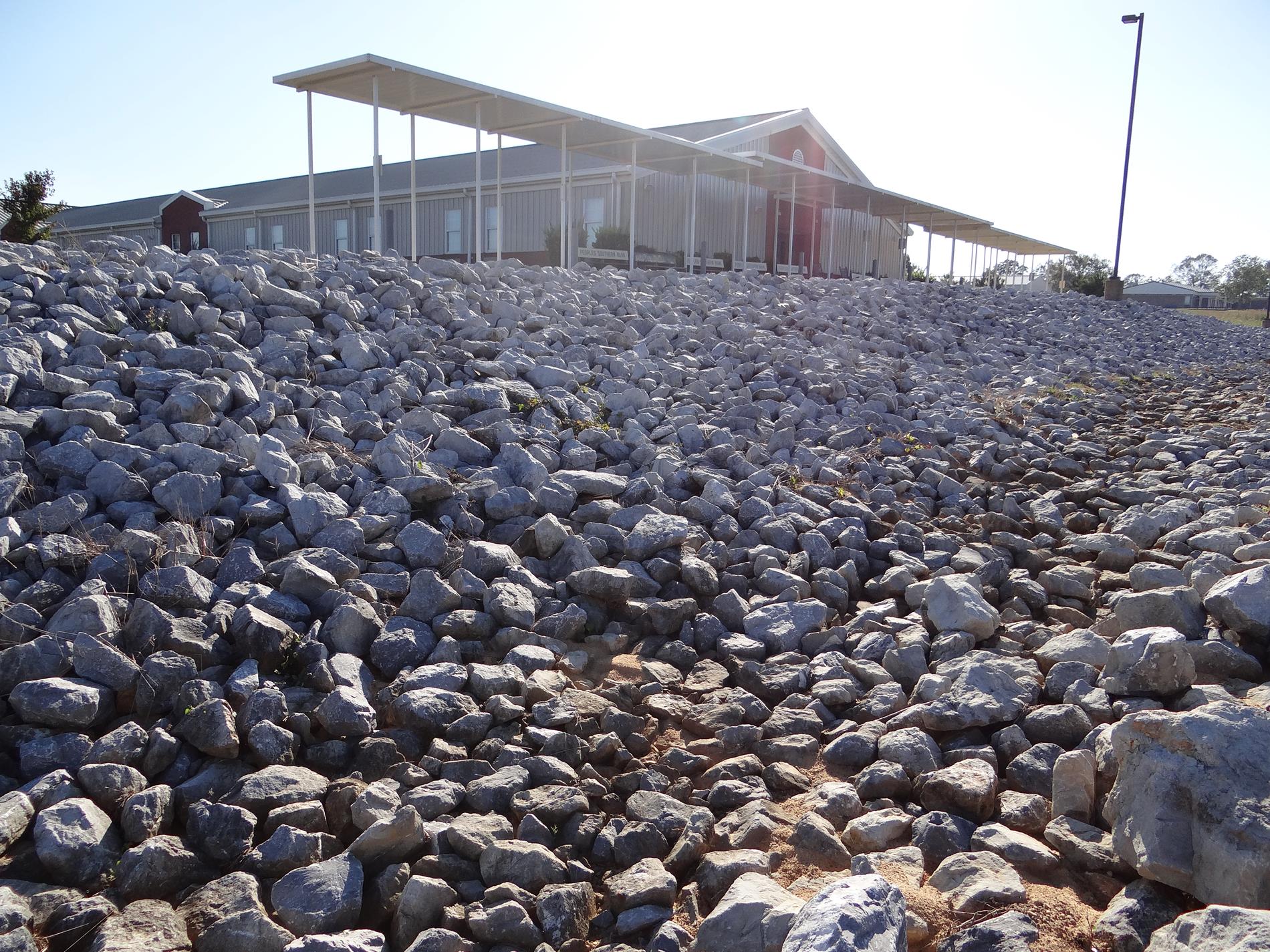 Angled View of the Bus-rider entrance of Clanton Middle School - A wall of large, white rocks form a barrier beside a long, white, metal awning covering a sidewalk that extends past the end of the building in either direction along the bus drop-off and pickup road