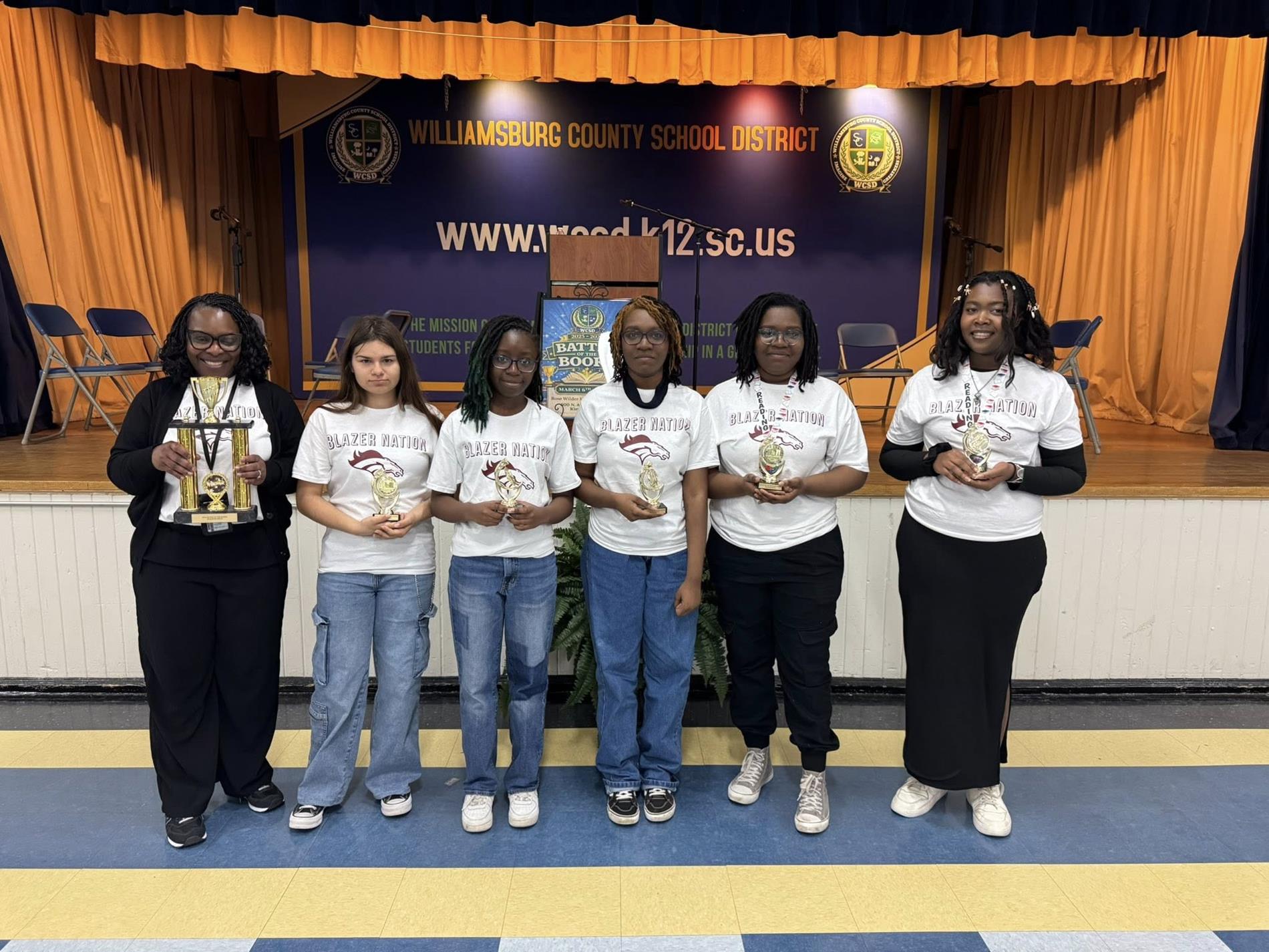 Six members of the Kingstree High School "Blazer Nation" team pose with trophies on a stage at the Williamsburg County School District auditorium. Five students wear matching white Blazer Nation t-shirts while standing in front of a purple district banner.