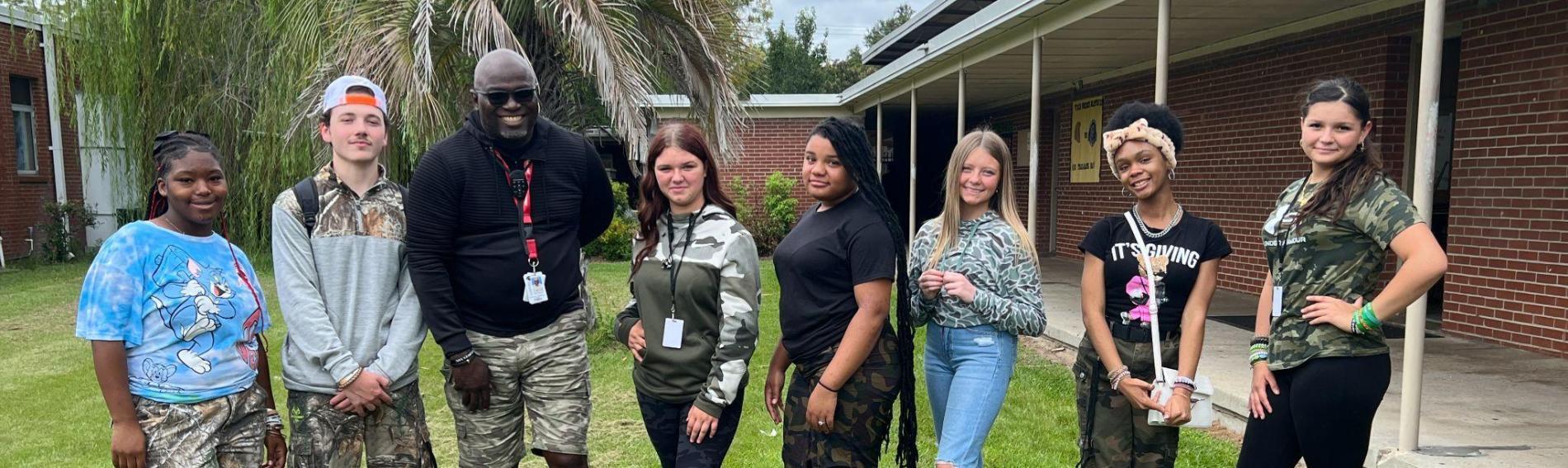 A teacher and seven students pose together in a line. They're all wearing camouflage for camo spirit day.  