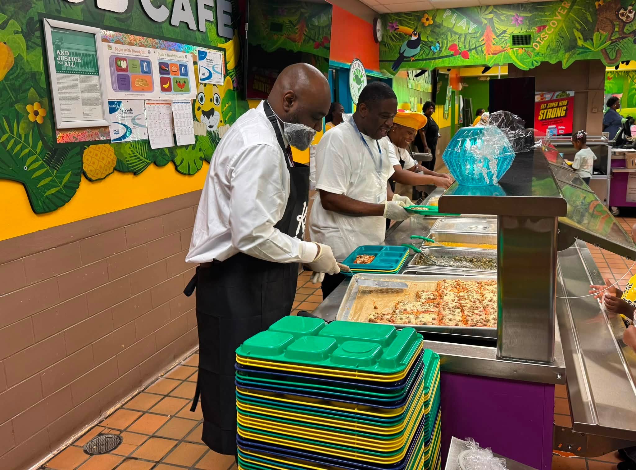 School cafeteria staff serve food to students along a lunch line in a brightly decorated dining area. Staff members wearing gloves and protective clothing prepare trays beside pans of pizza and other foods, with stacks of colorful lunch trays in the foreground.