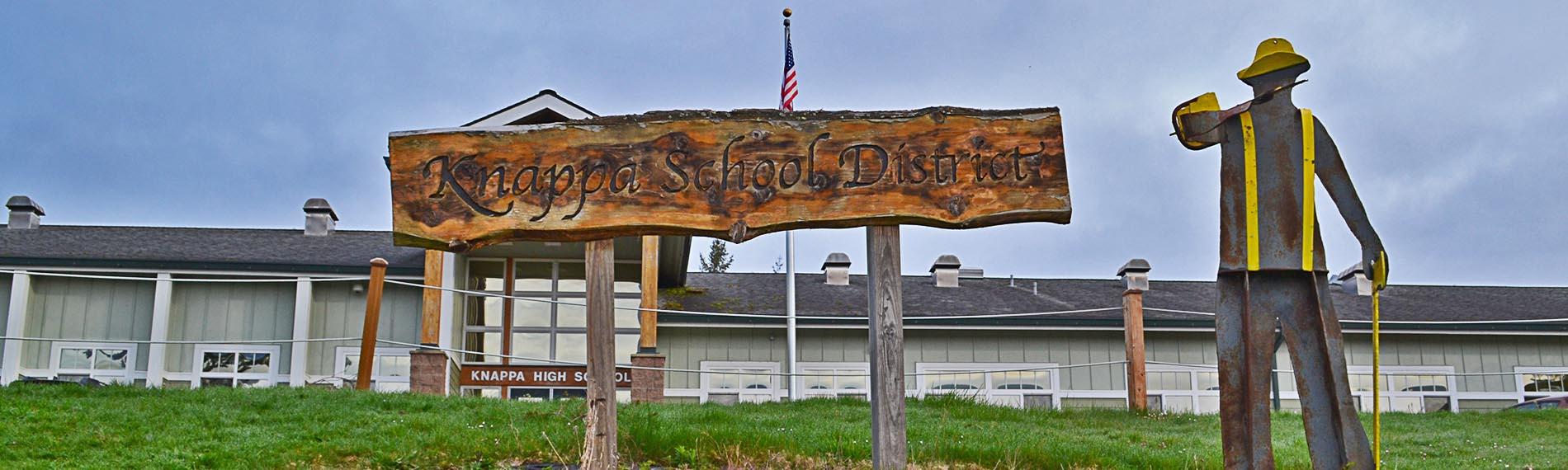 Sign reading "Knappa School District" outside the high school and logger statue