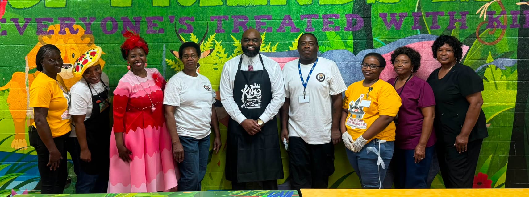 Nine school staff members stand together in front of a brightly colored cafeteria mural. The group wears a mix of school-themed T-shirts, kitchen attire, colorful clothing, and medical-style scrubs. At the center, a man in a white shirt and tie wears a black “King of the Kitchen” apron.