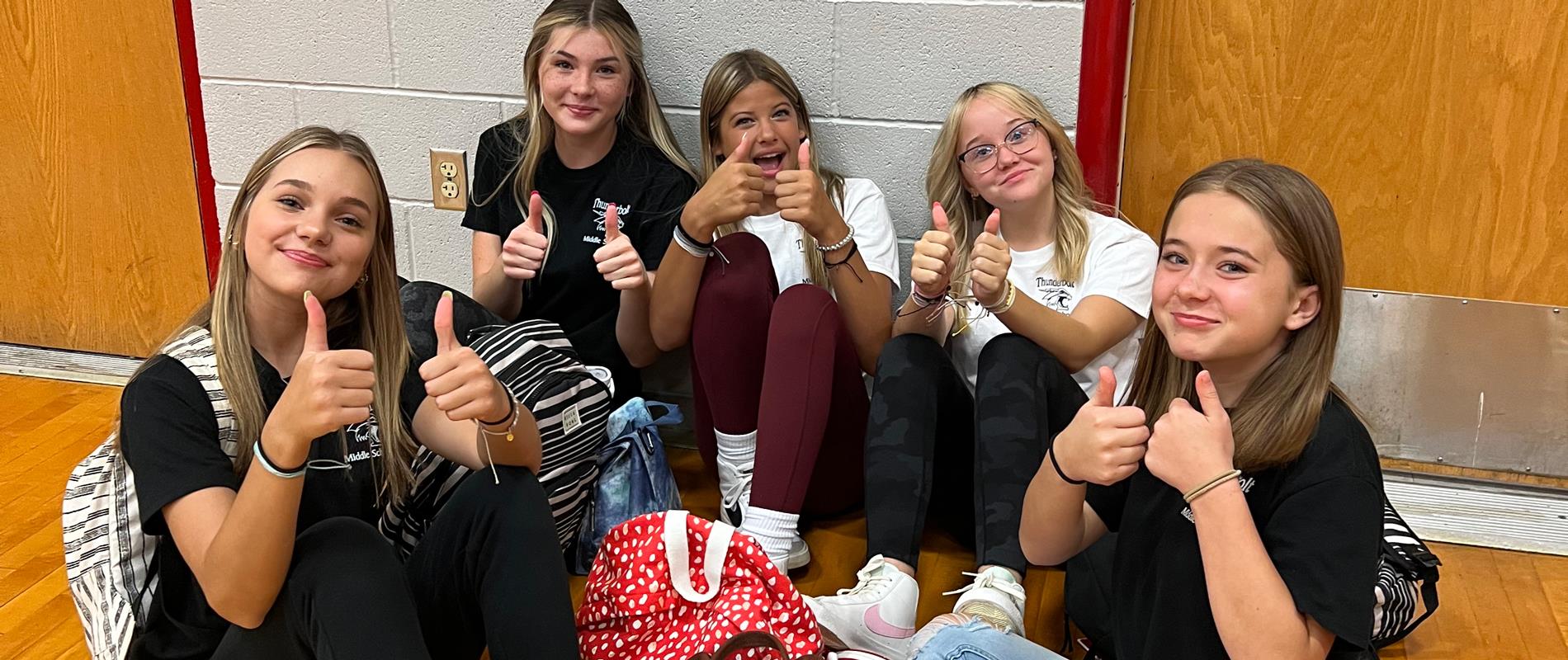 Group of girls sitting in the gym giving a thumbs up