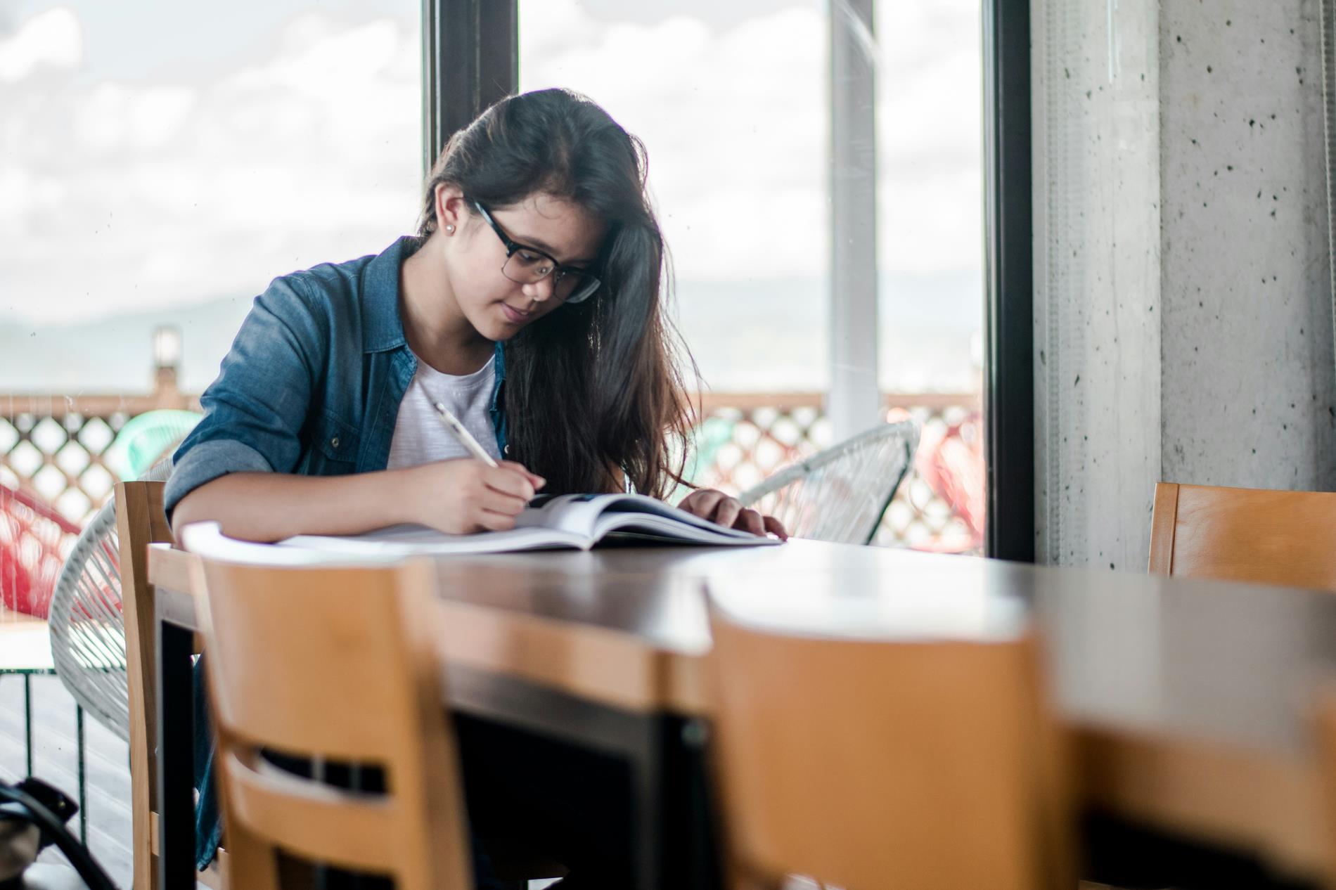 A young woman sits at a table with a pencil studying and writing in a book