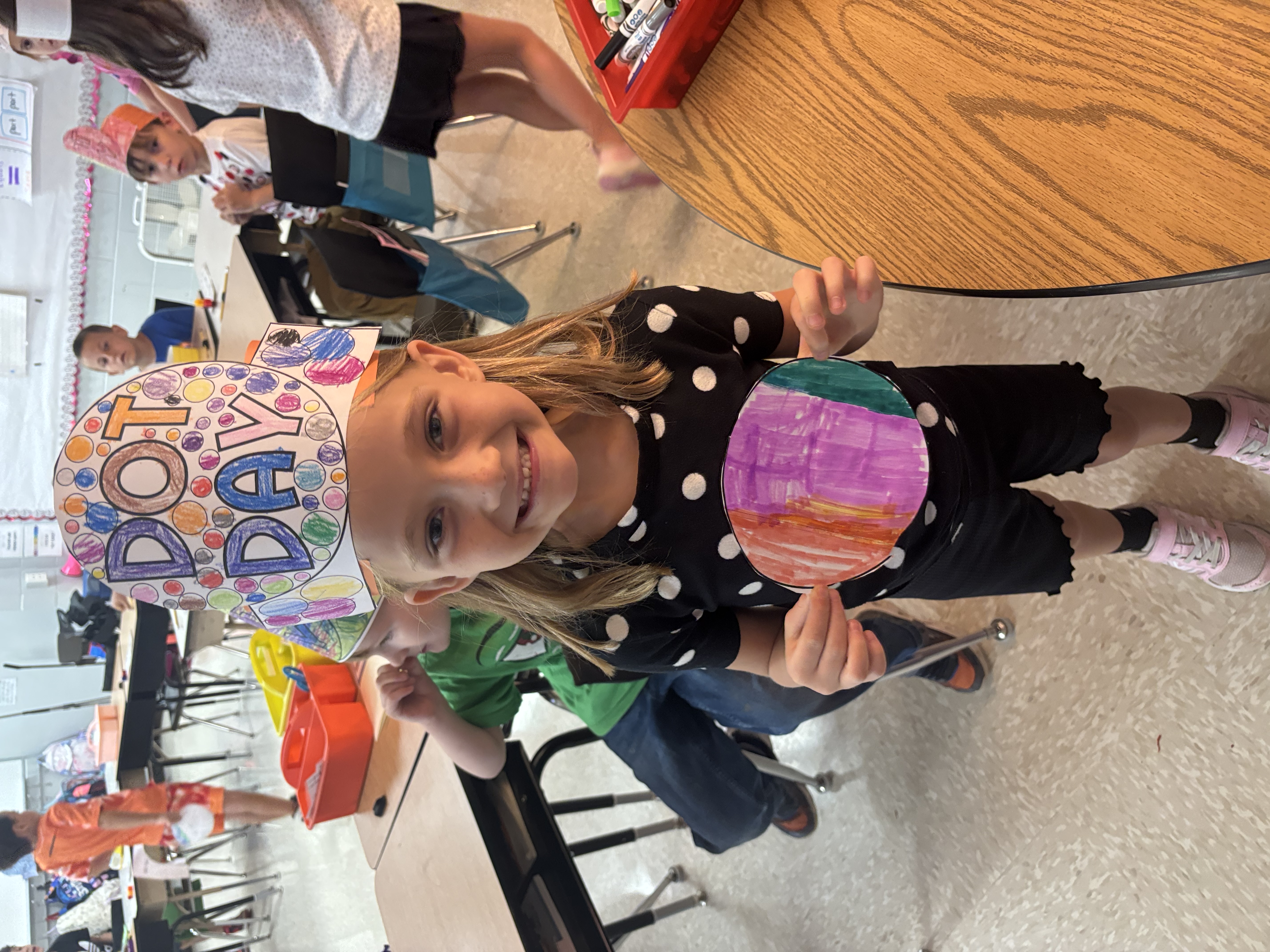 female student holding a dot she colored while wearing a hand-made hat that says dot day