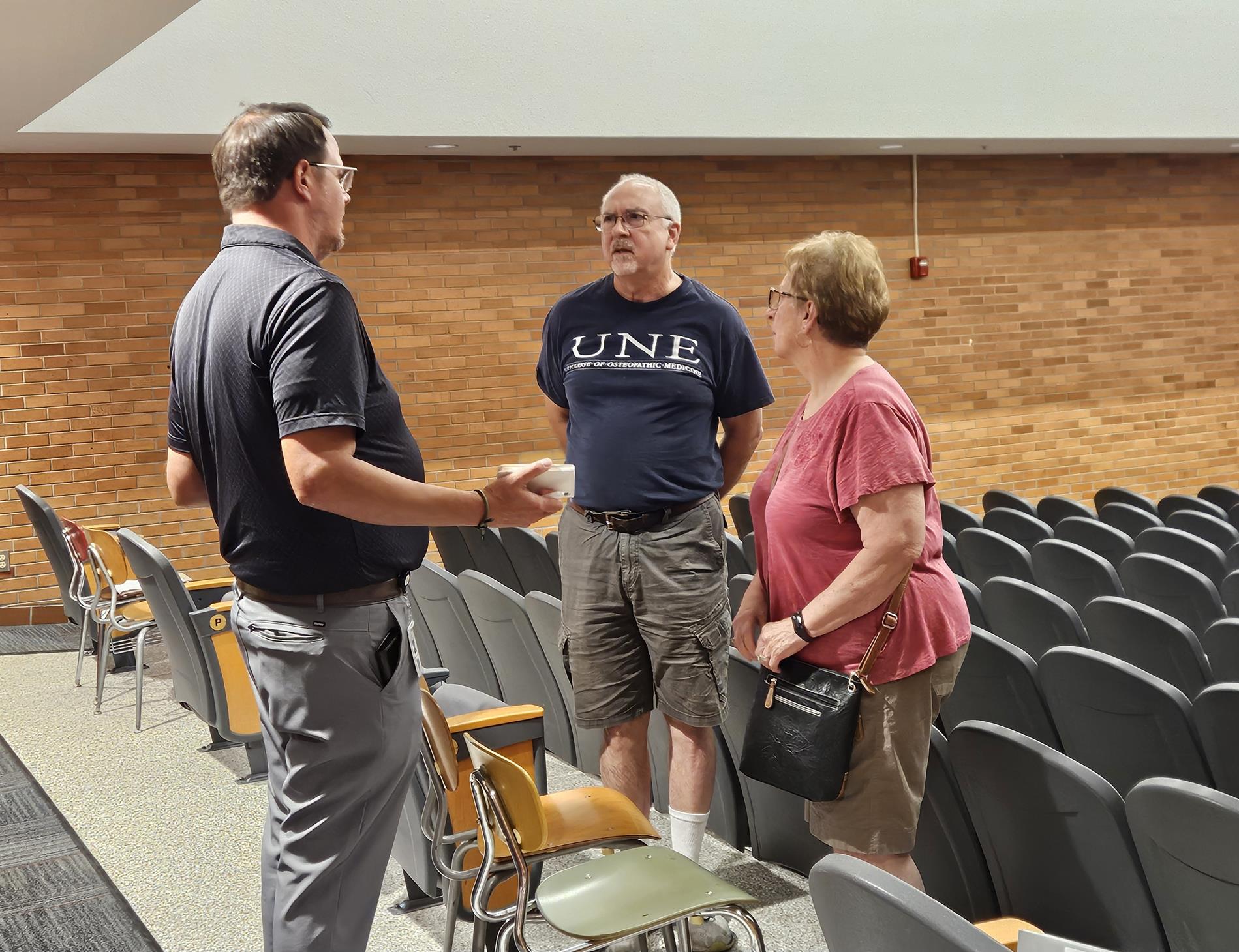 Two men and a women stand at the back of the auditorium chatting about the presentation