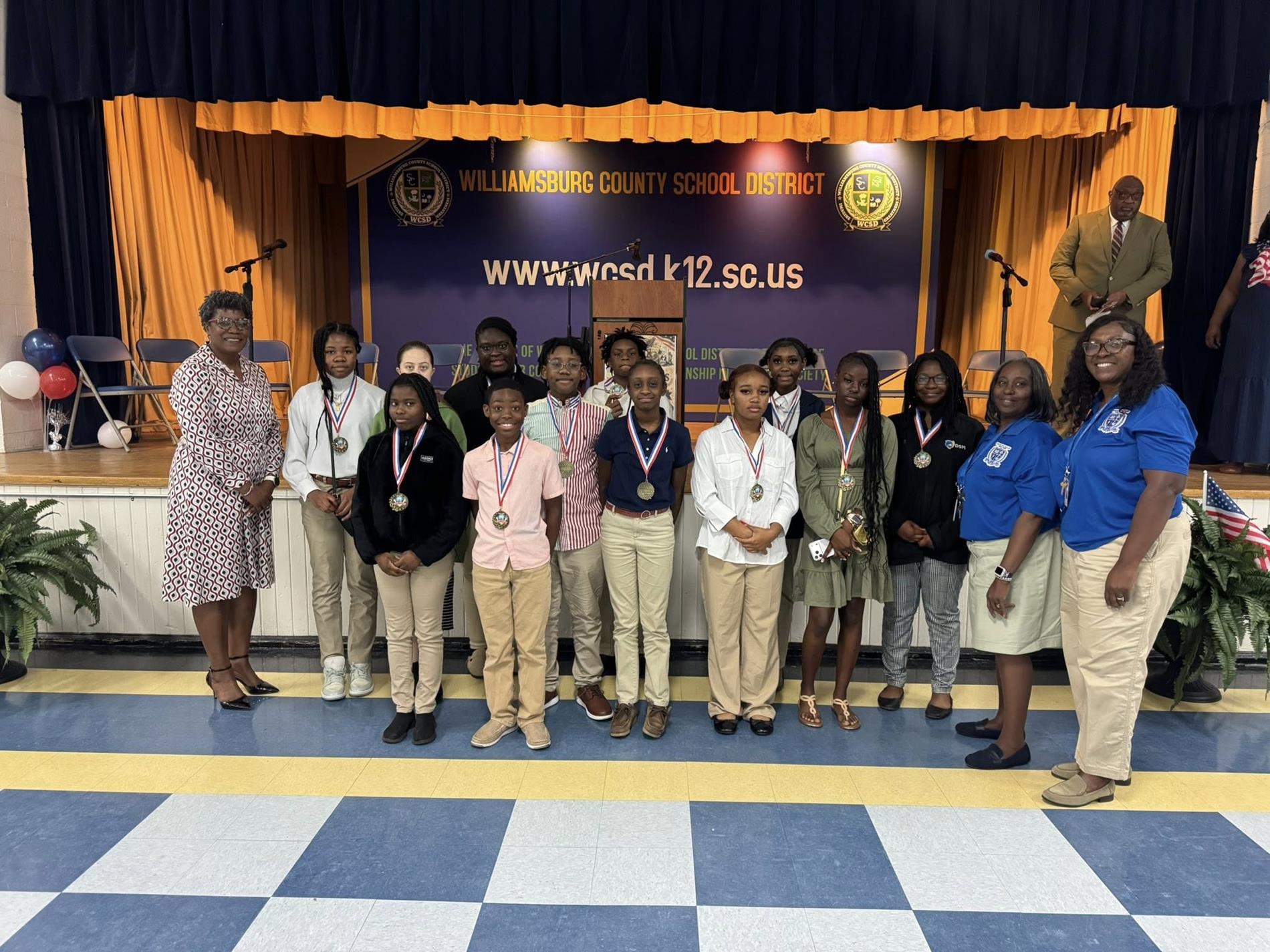 A group of middle school students stand in front of a stage with a “Williamsburg County School District” banner and website displayed behind them. The students wear school uniforms or casual outfits and have medals around their necks, posing in a line. Several adults stand alongside them, one holding a trophy. The stage features gold curtains, chairs, microphones, and a podium, indicating an awards or recognition ceremony.