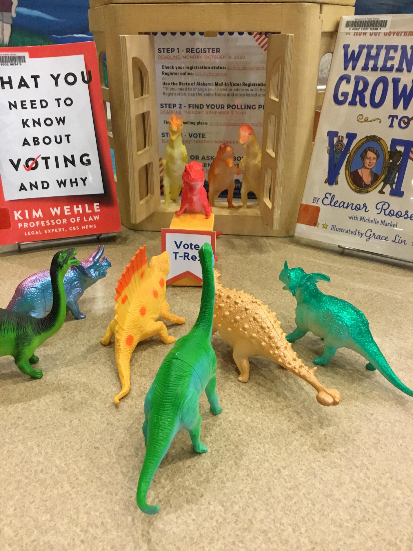 Dinosaur toy figures gathered around a toy figure t-rex standing at a toy podium with a banner reading "VOTE T-Rex" surrounded by books about voting