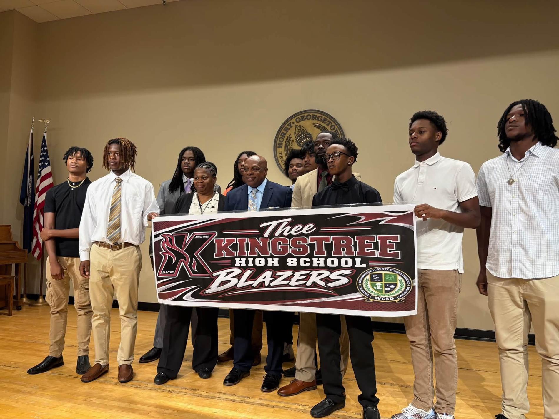 Kingstree High School students and adults pose on a stage holding a large “Thee Kingstree High School Blazers” banner.
