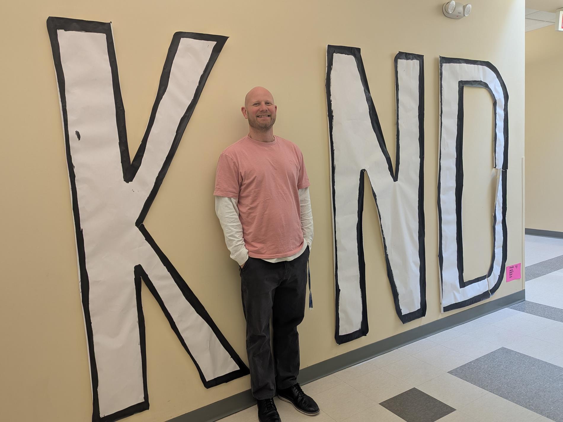 Picture of smiling bald man wearing a pink shirt. The man stands agains a the wall, forming the "i" in a large sign reading "KIND".