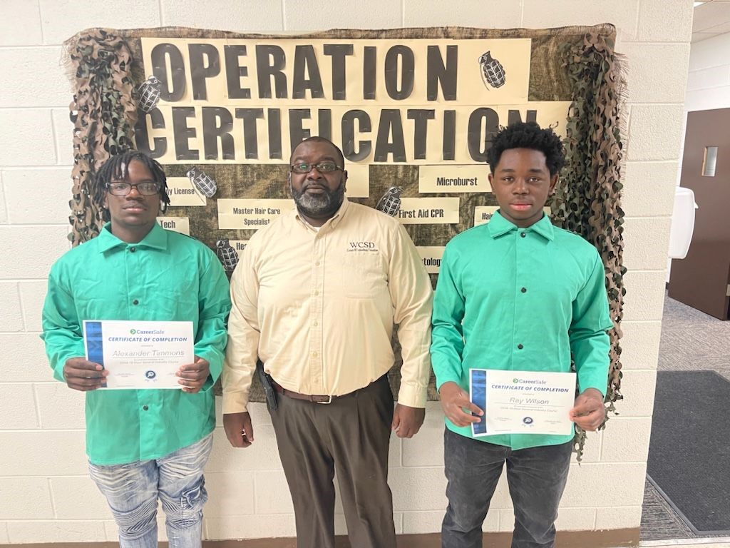 operation certification. two young men along with male principal standing in front of bulletin board holding award certificates. 