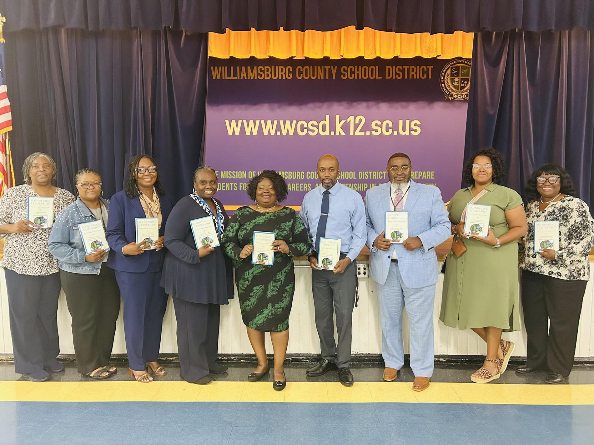 A group of nine adults stand in a row holding matching books in front of a stage backdrop that reads "Williamsburg County School District" and "www.wcsd.k12.sc.us