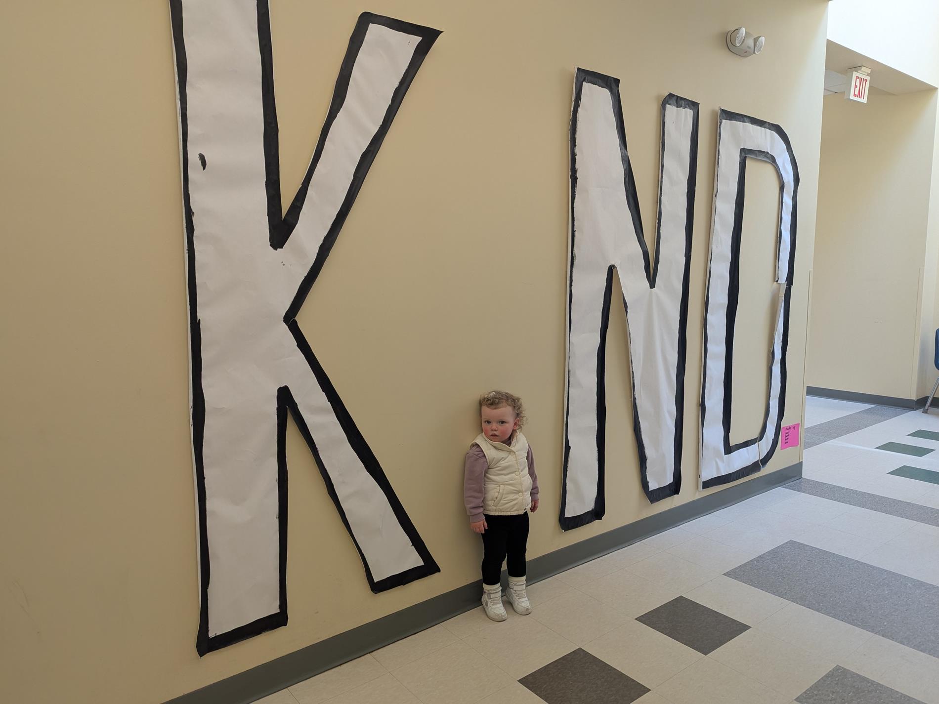 Picture of assistant coordinator of kindness wearing a pink shirt. The assistant coordinator of kindness stands agains a the wall, forming the "i" in a large sign reading "KIND".