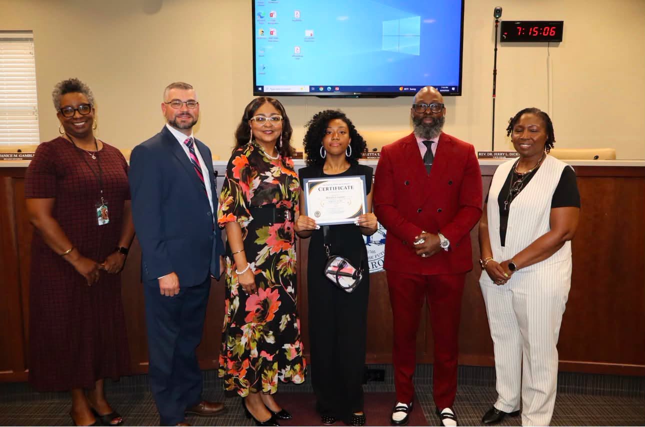 A student stands at the center of a group of five adults while holding a certificate of recognition. The group poses together in a school board meeting room, with a large display screen and board seating visible in the background.