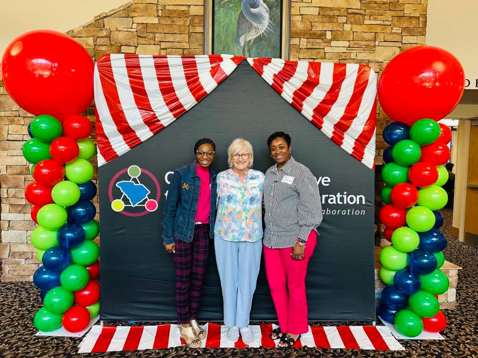 Three women stand together smiling in front of a decorative event backdrop. The backdrop features red-and-white striped draping resembling a carnival tent and colorful balloon columns topped with large red balloons on each side. The women pose shoulder to shoulder in a lobby or conference space with a stone wall behind them. The setting appears festive and welcoming, suggesting a community or educational event.