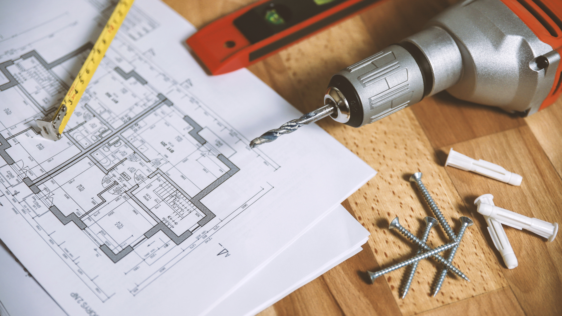 A building blueprint with a drill and a tape measure and nails sit on a wooden table
